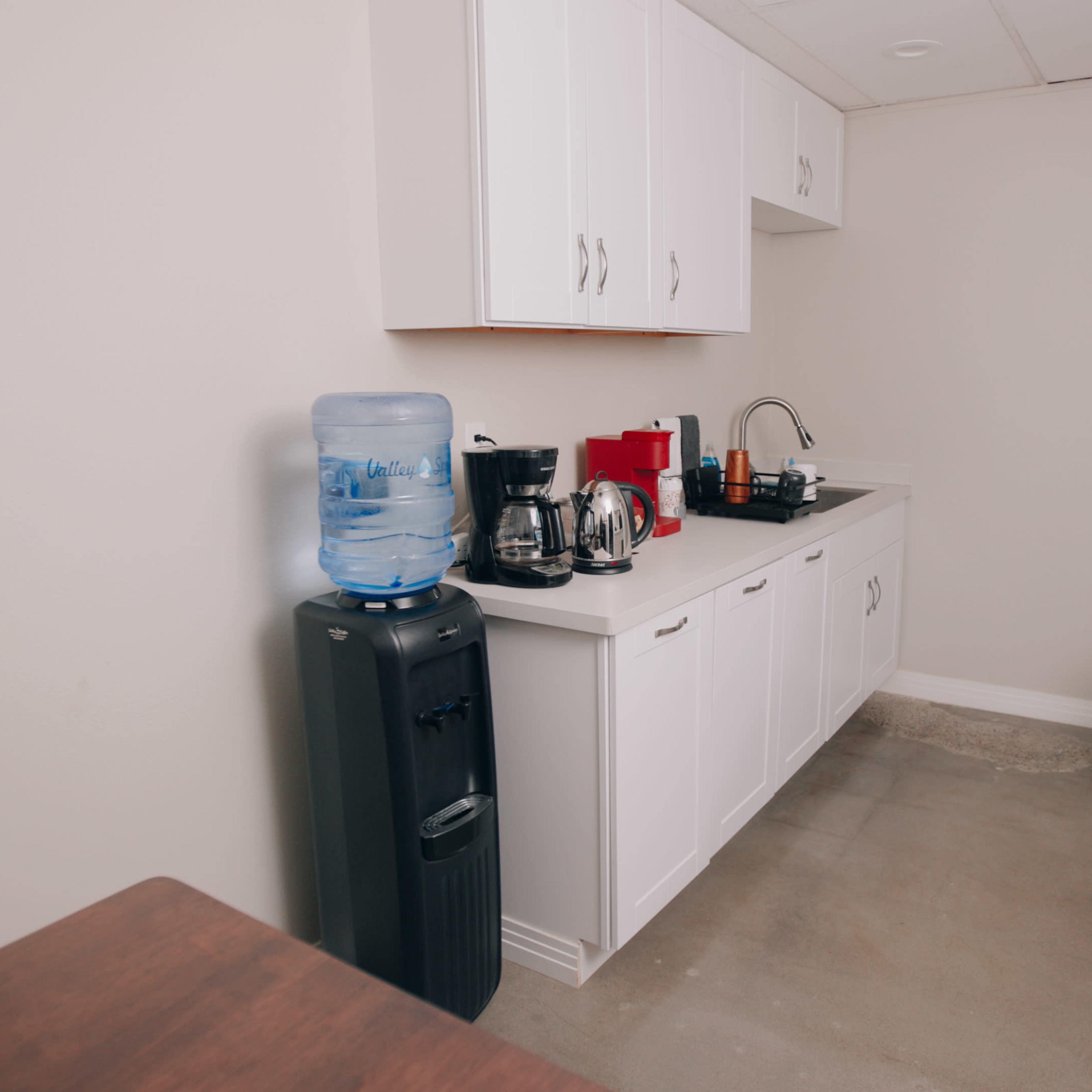 A kitchen with white cabinets, a water cooler, coffee maker, and a refrigerator along the wall.