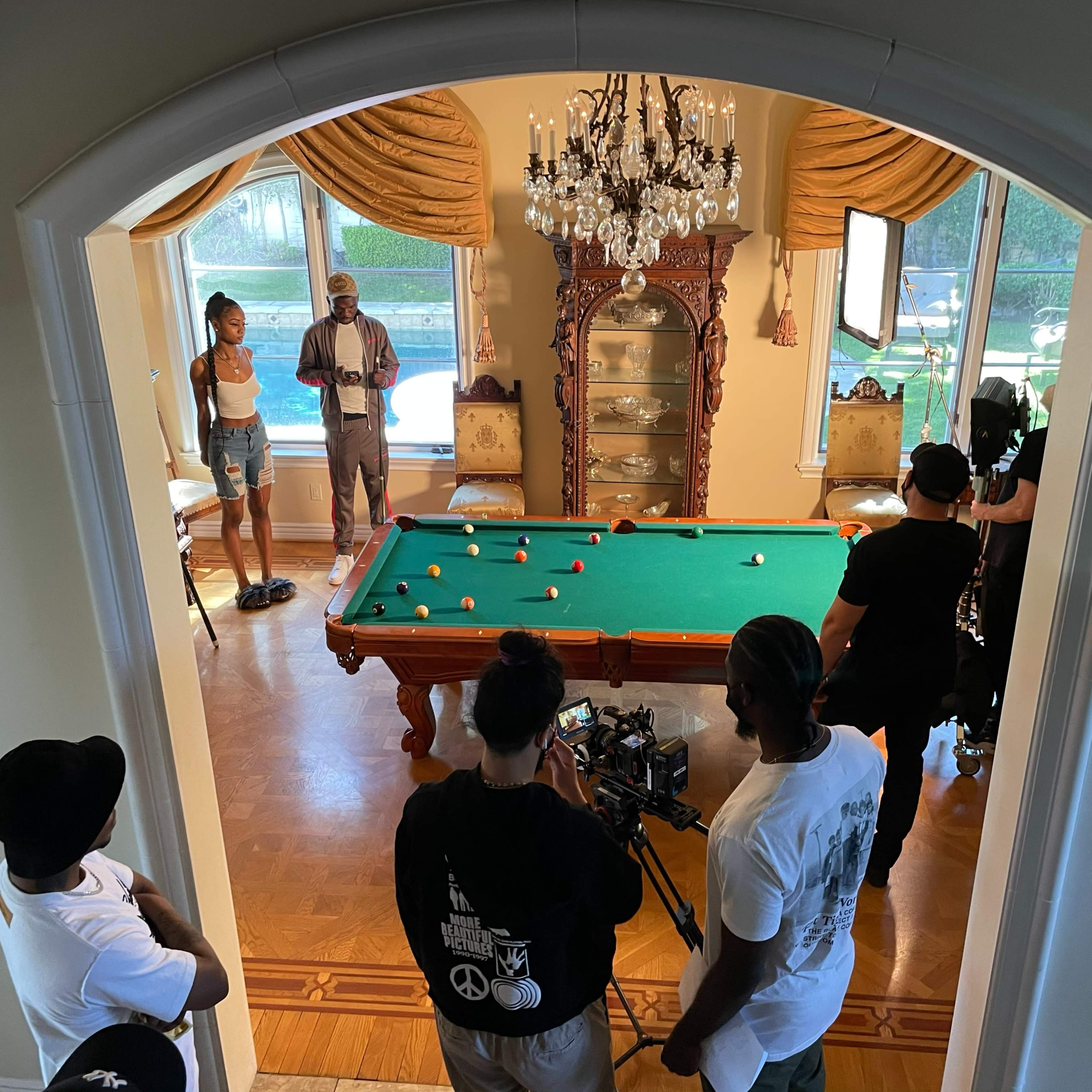 A film crew is shooting a scene involving a pool table in a well-lit room with large windows and elegant furniture.