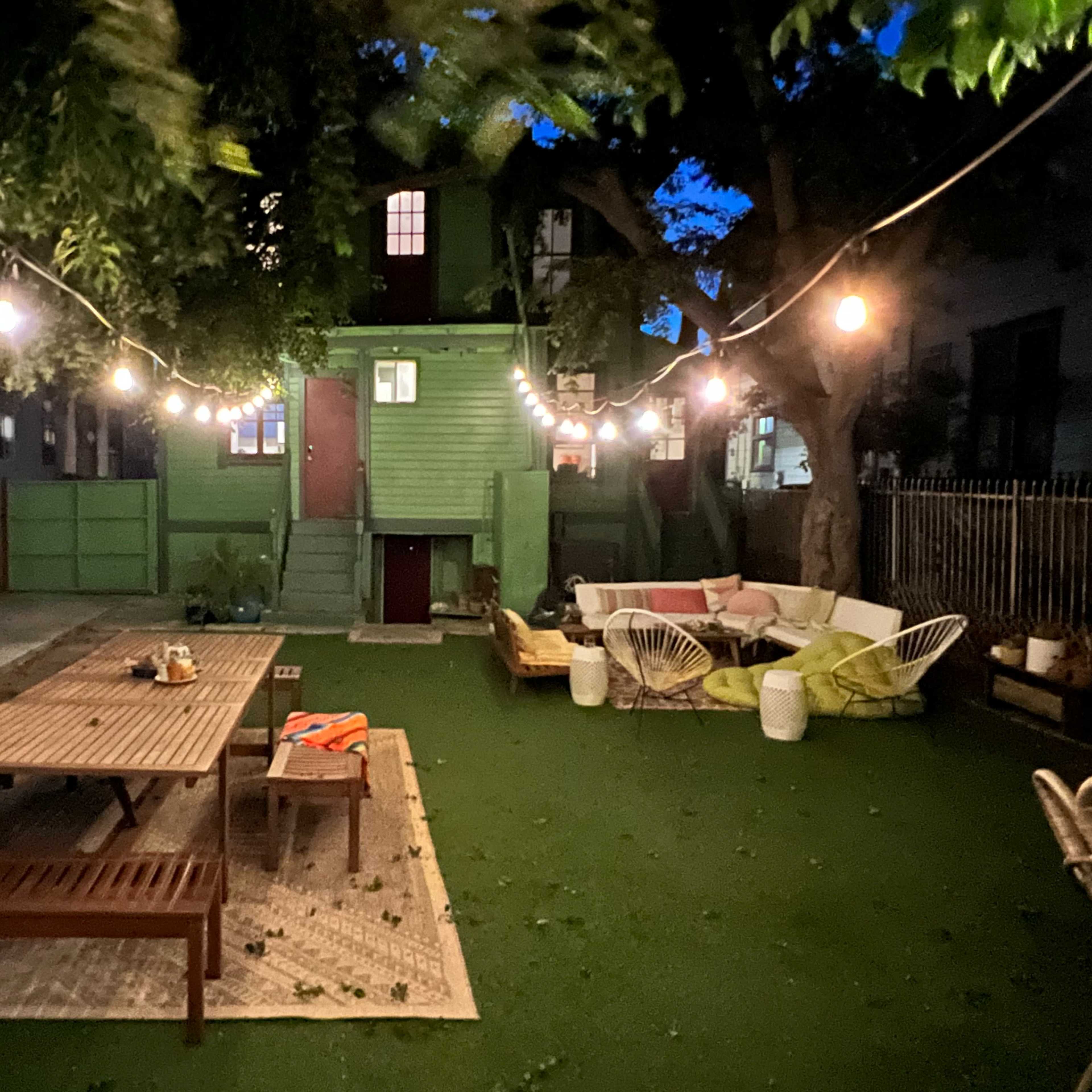 A green two-story building in a backyard with a wooden dining table, lounge chairs, and string lights illuminating the evening scene.