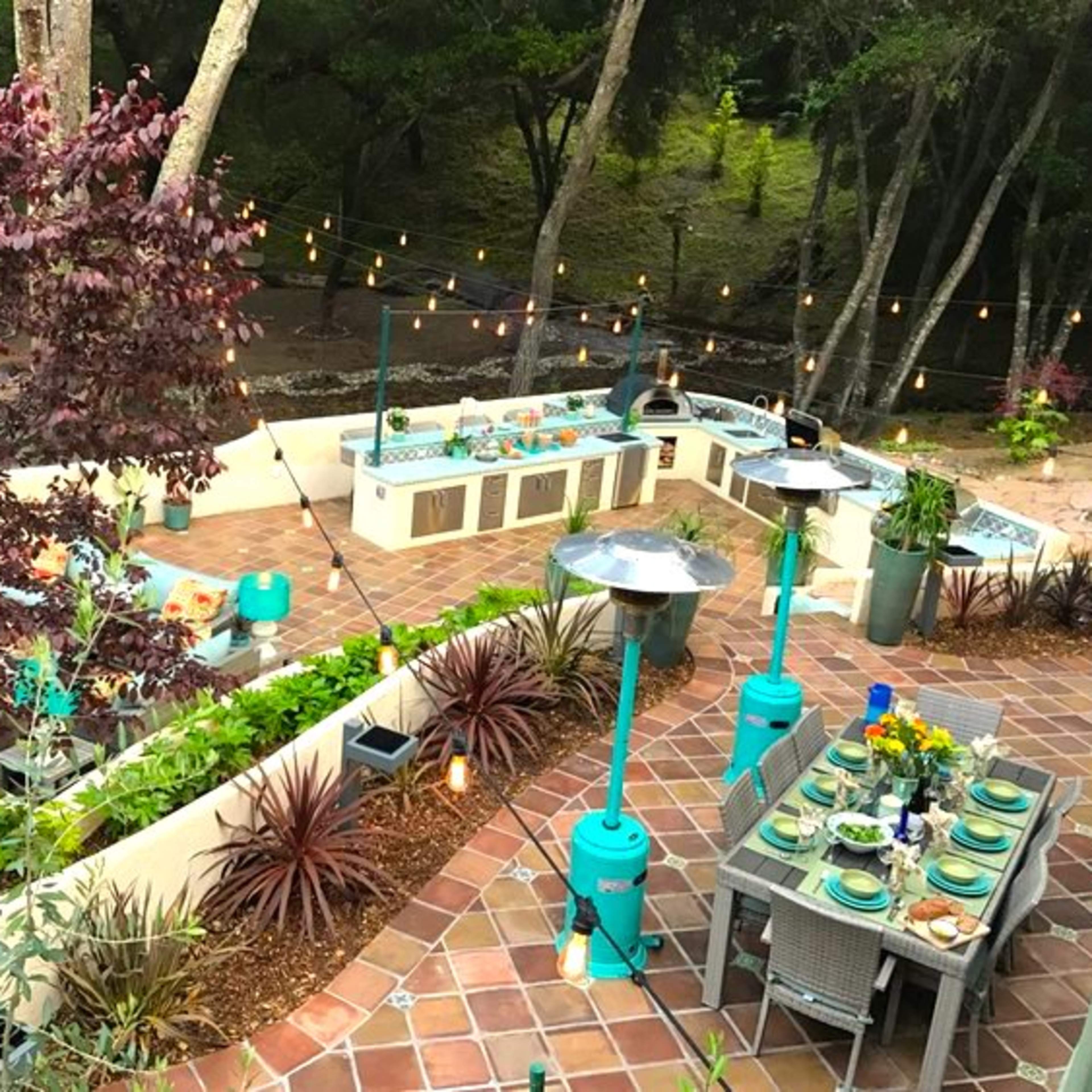 An outdoor kitchen and dining area decorated with string lights, surrounded by trees and various plants.