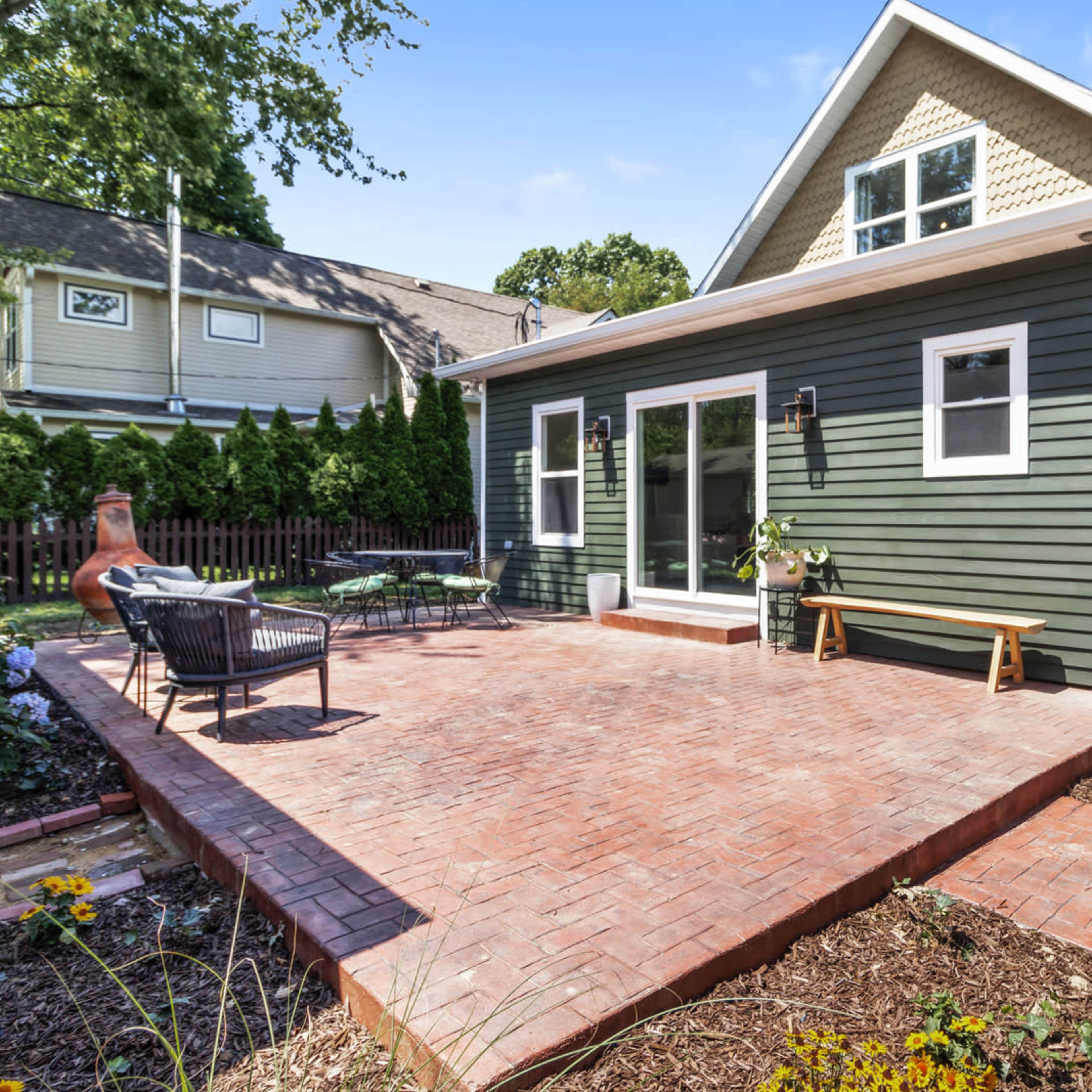 A green house with a brick patio and a wooden privacy screen, surrounded by landscaped gardens and neighboring homes.