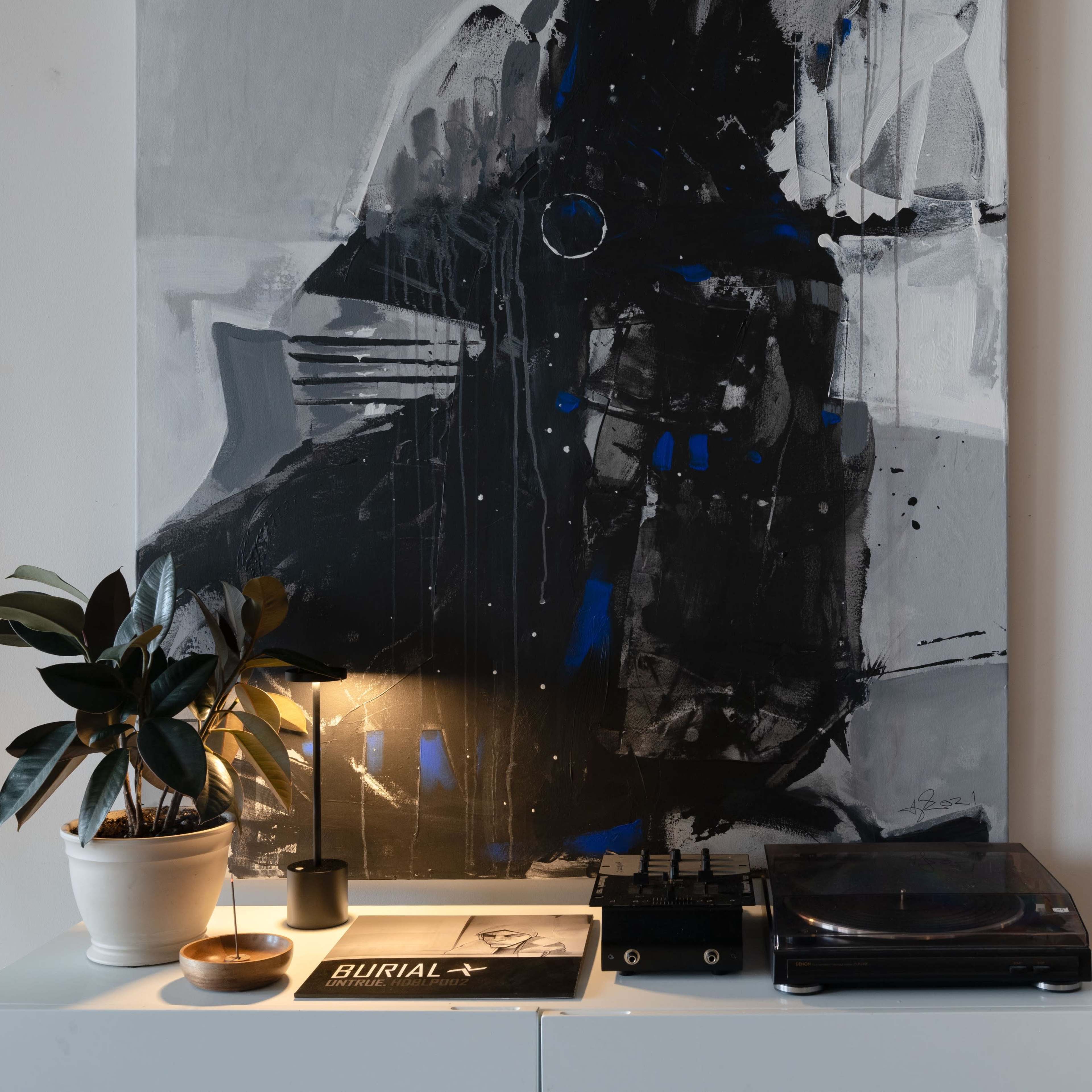A modern white cabinet holds a turntable and records beneath a large abstract painting, accompanied by a small lamp and a potted plant.