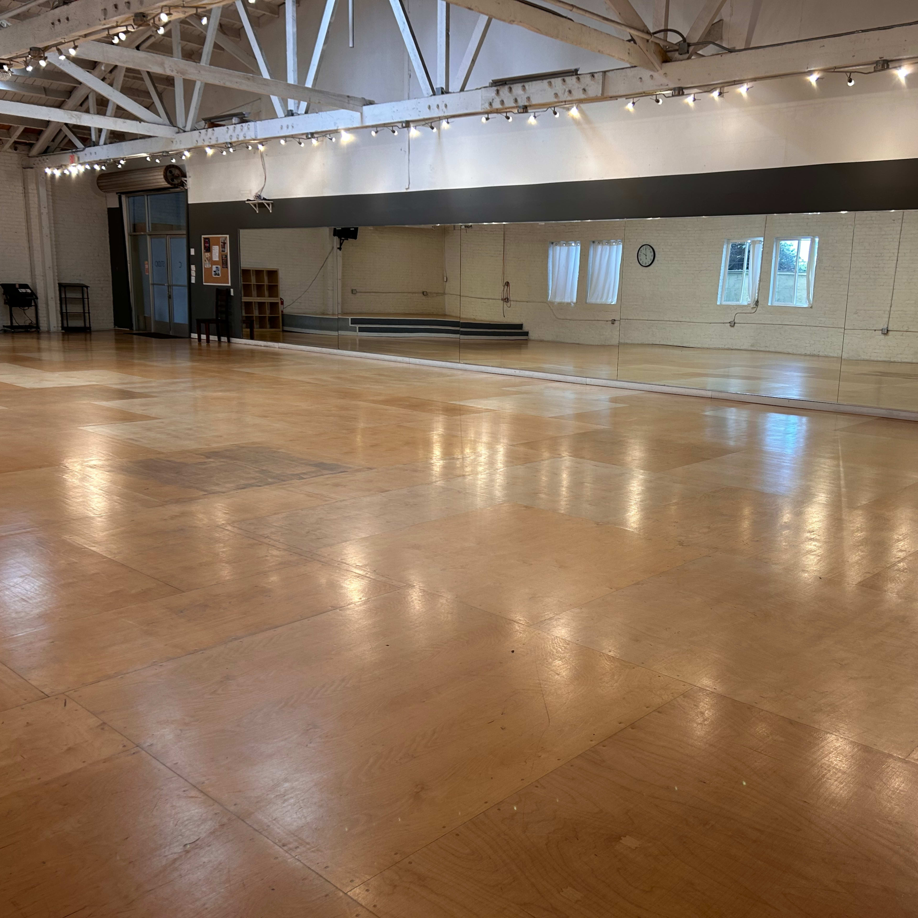 A spacious dance studio with a polished wooden floor, large mirrors along one wall, and natural light coming through several windows.