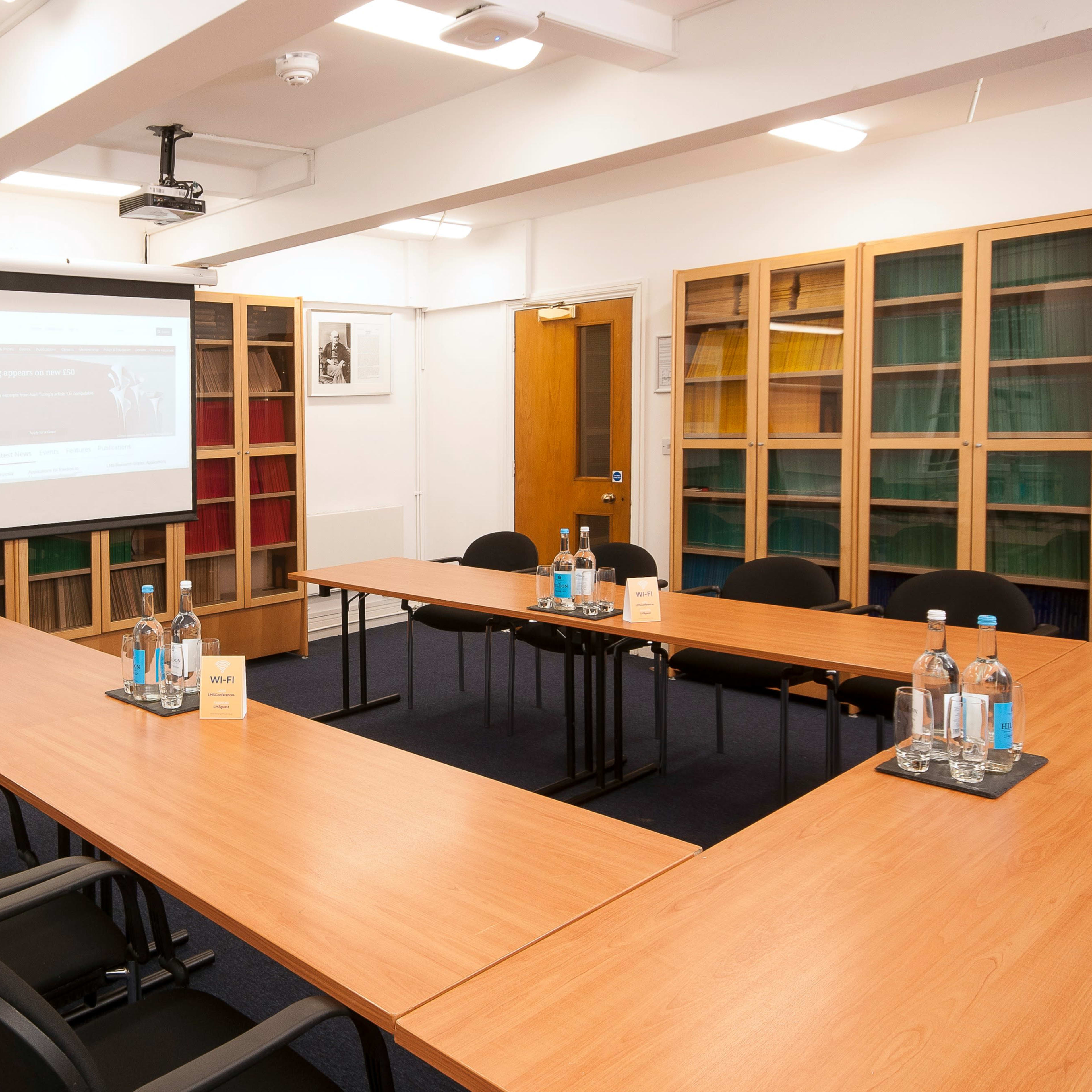 A conference room features a large rectangular table surrounded by black chairs, a projector screen at the front, and shelves filled with books in the background.