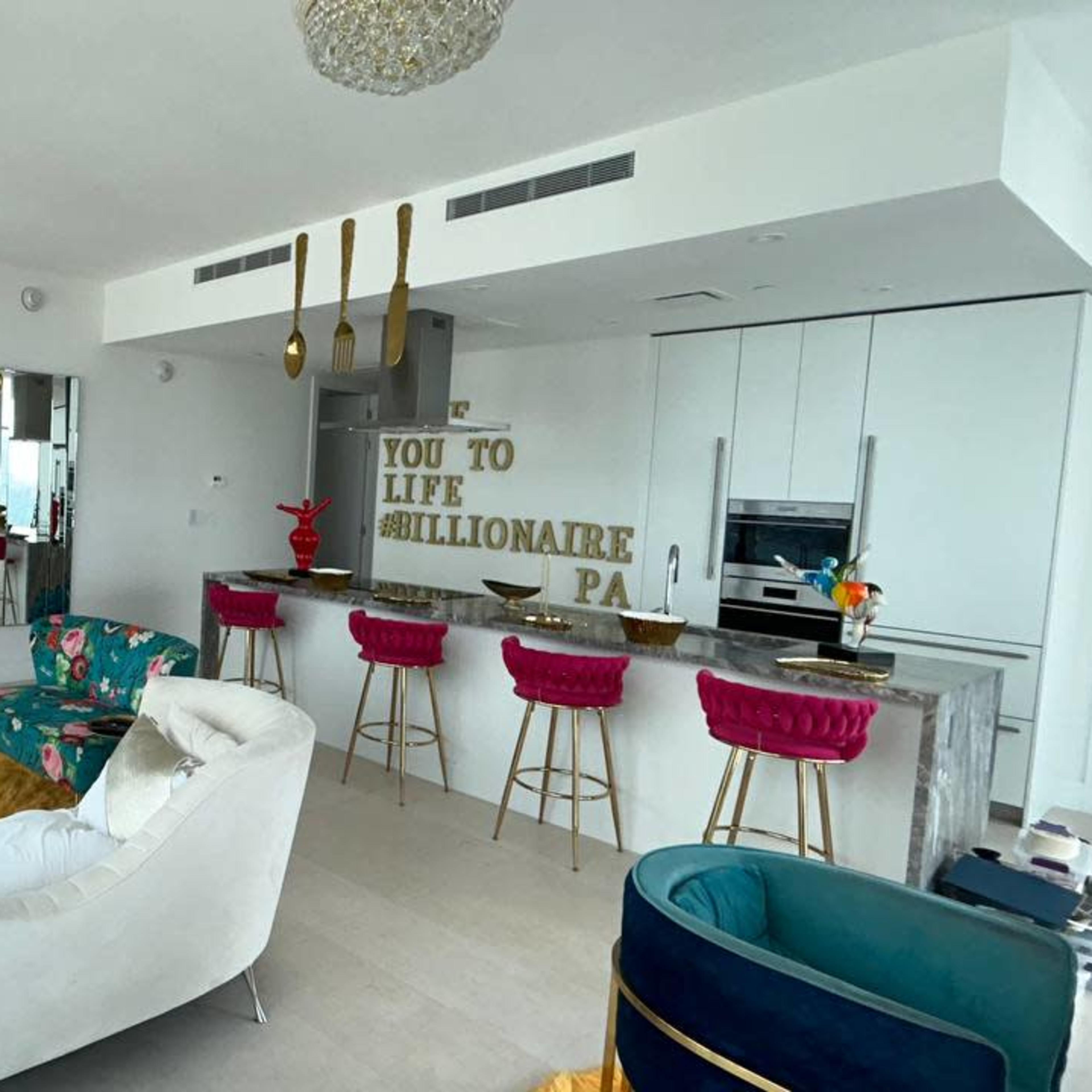 A modern, stylish living room features a large television, a plush white sofa, and a kitchen bar with colorful stools, adorned with decorative items and vibrant furnishings.