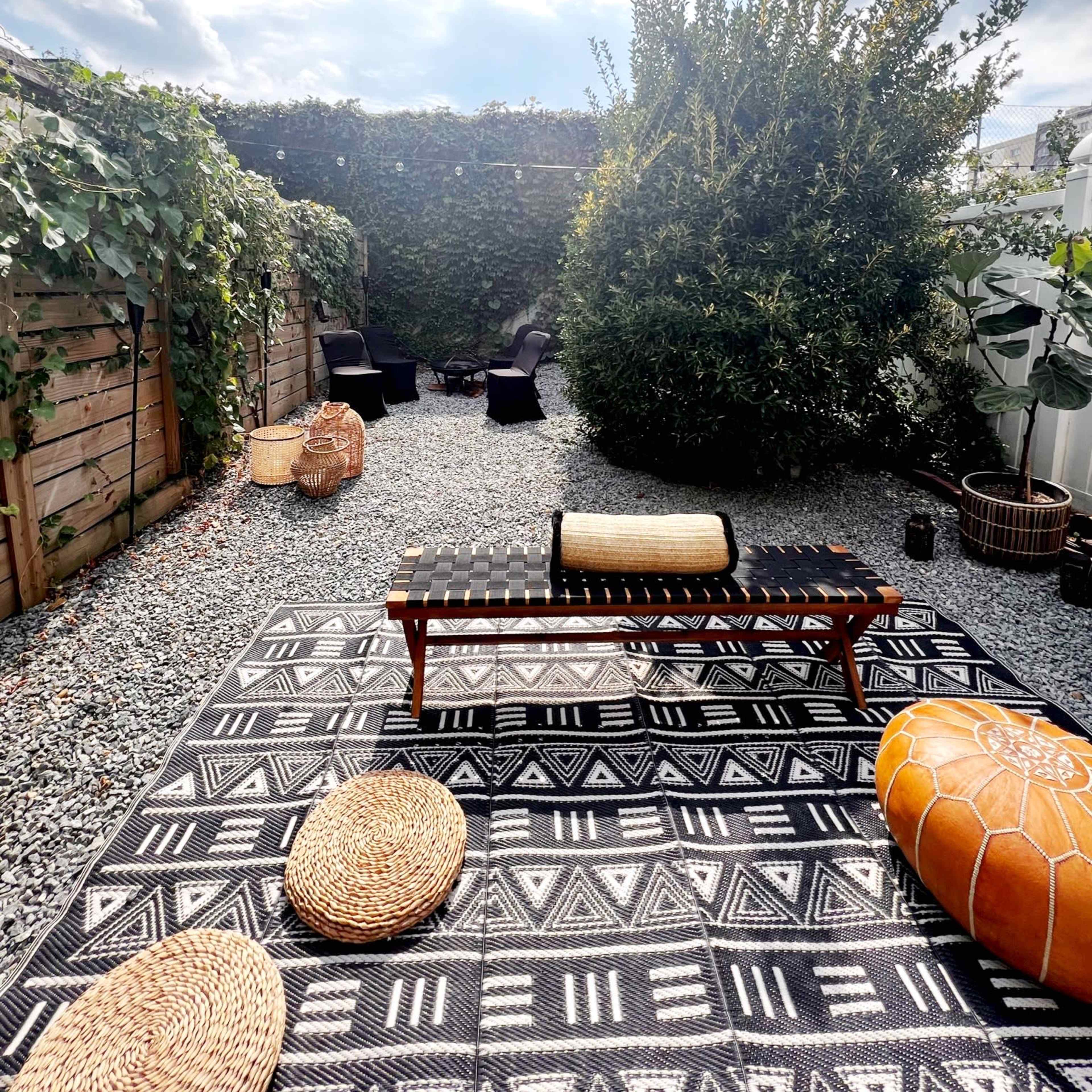 A gravel outdoor space with a patterned rug, a wooden bench, and various seating arrangements surrounded by greenery and string lights above.