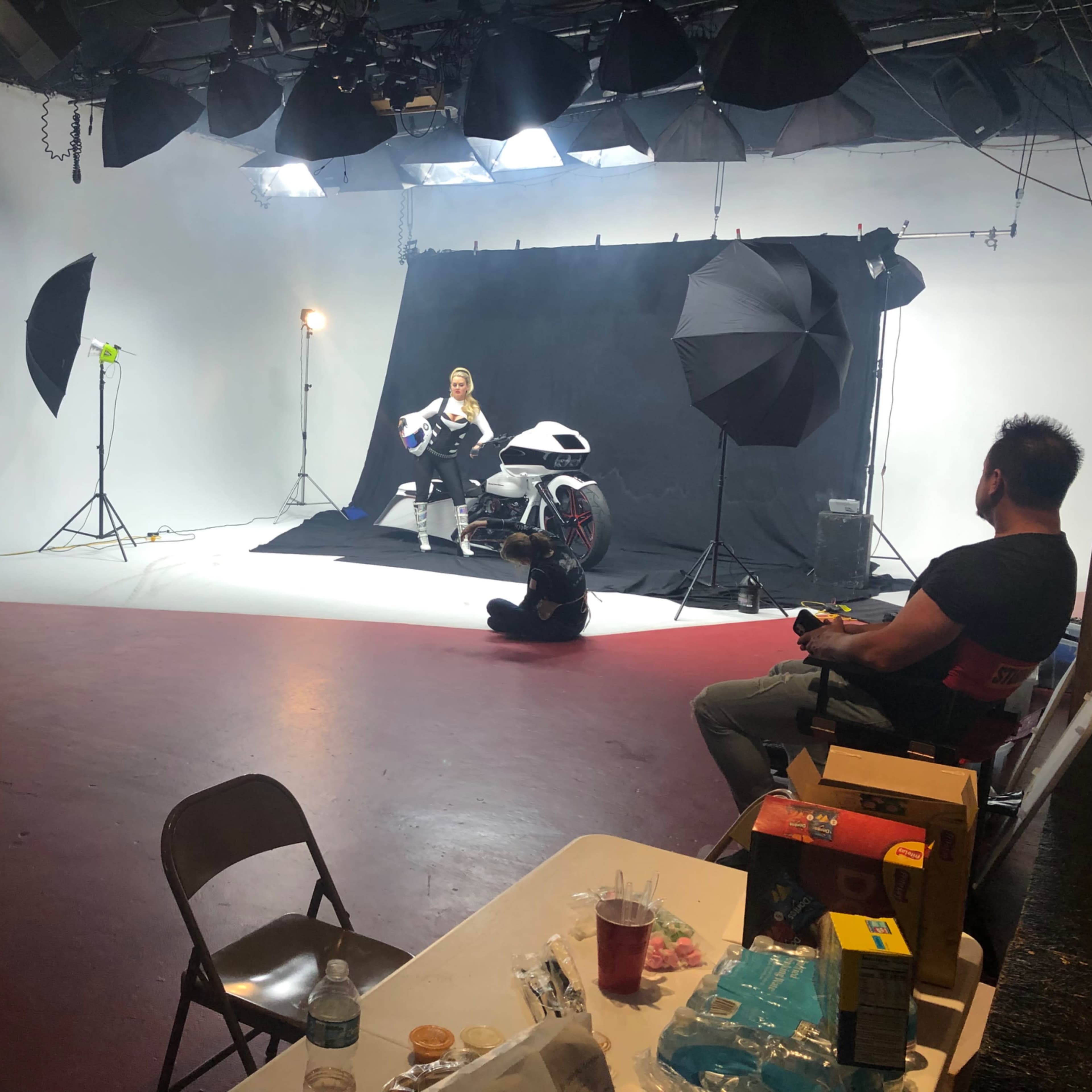 A photoshoot is underway in a studio with a model posing on a motorcycle, while crew members work in the background.