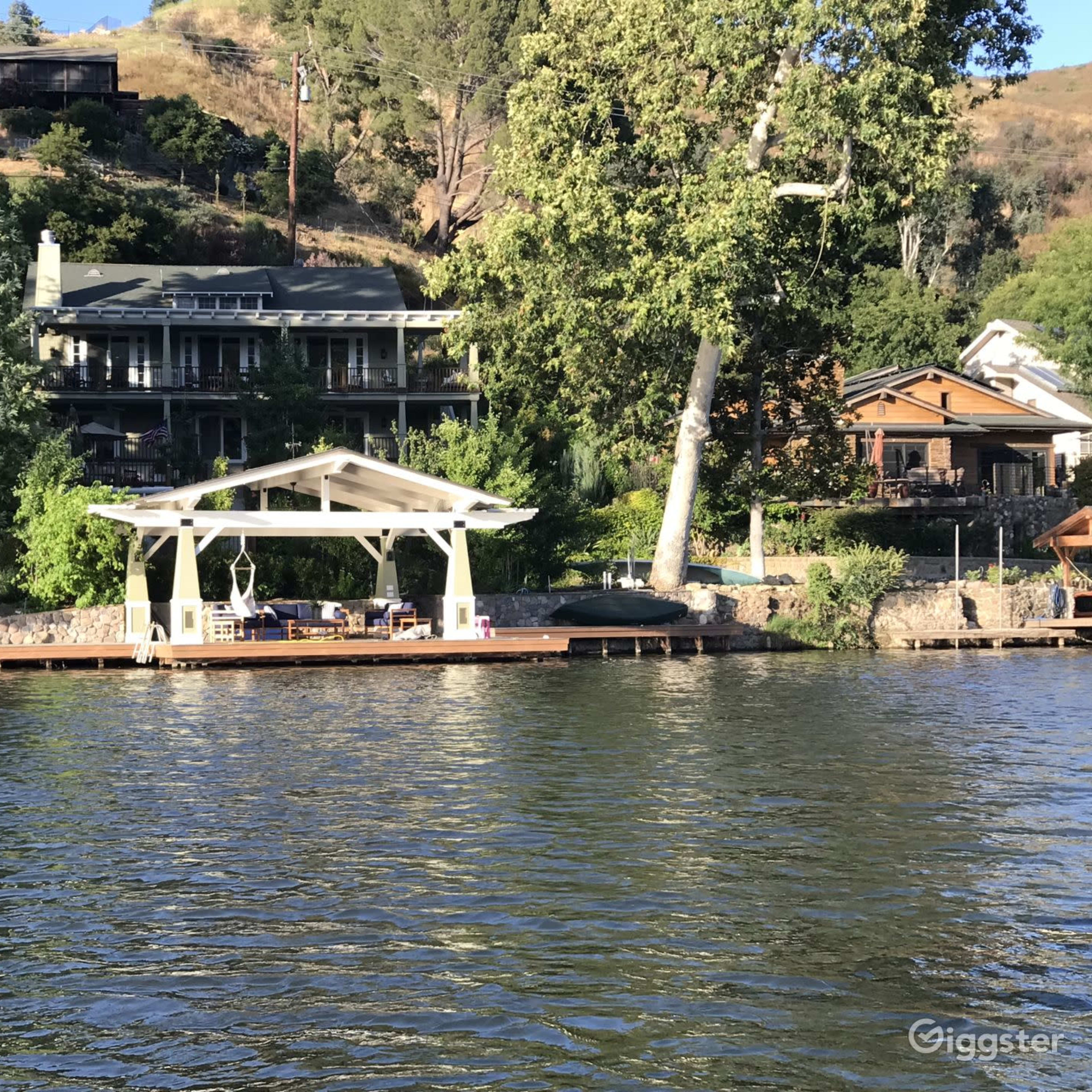 A waterfront scene with a dock featuring a covered seating area and several houses nestled on a hill behind it.