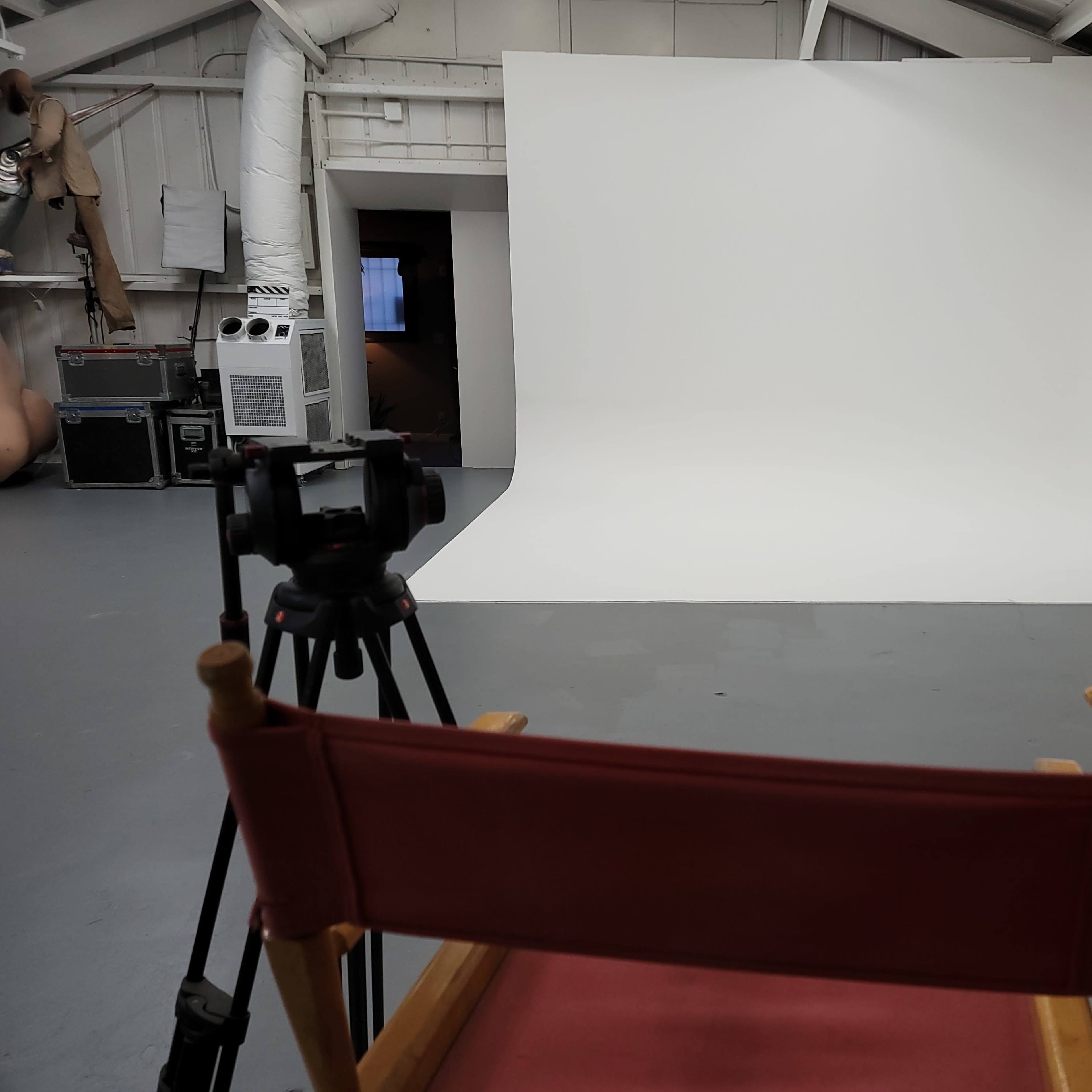 A photography studio features a white backdrop, a tripod-mounted camera, and two red director's chairs facing the scene.