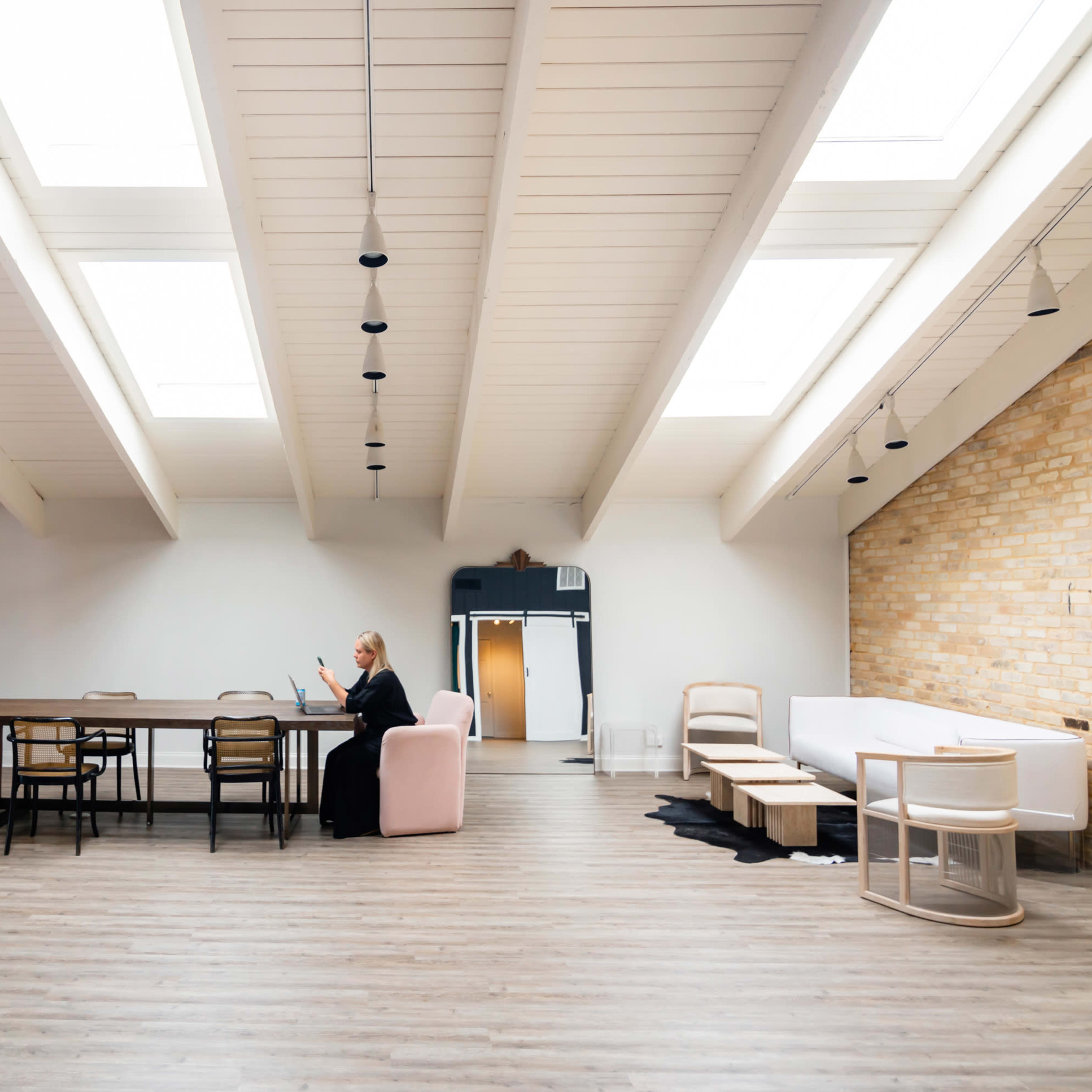 A spacious interior with a long wooden table, several chairs, and a woman sitting on a pink armchair, surrounded by large skylights and a mix of brick and painted walls.