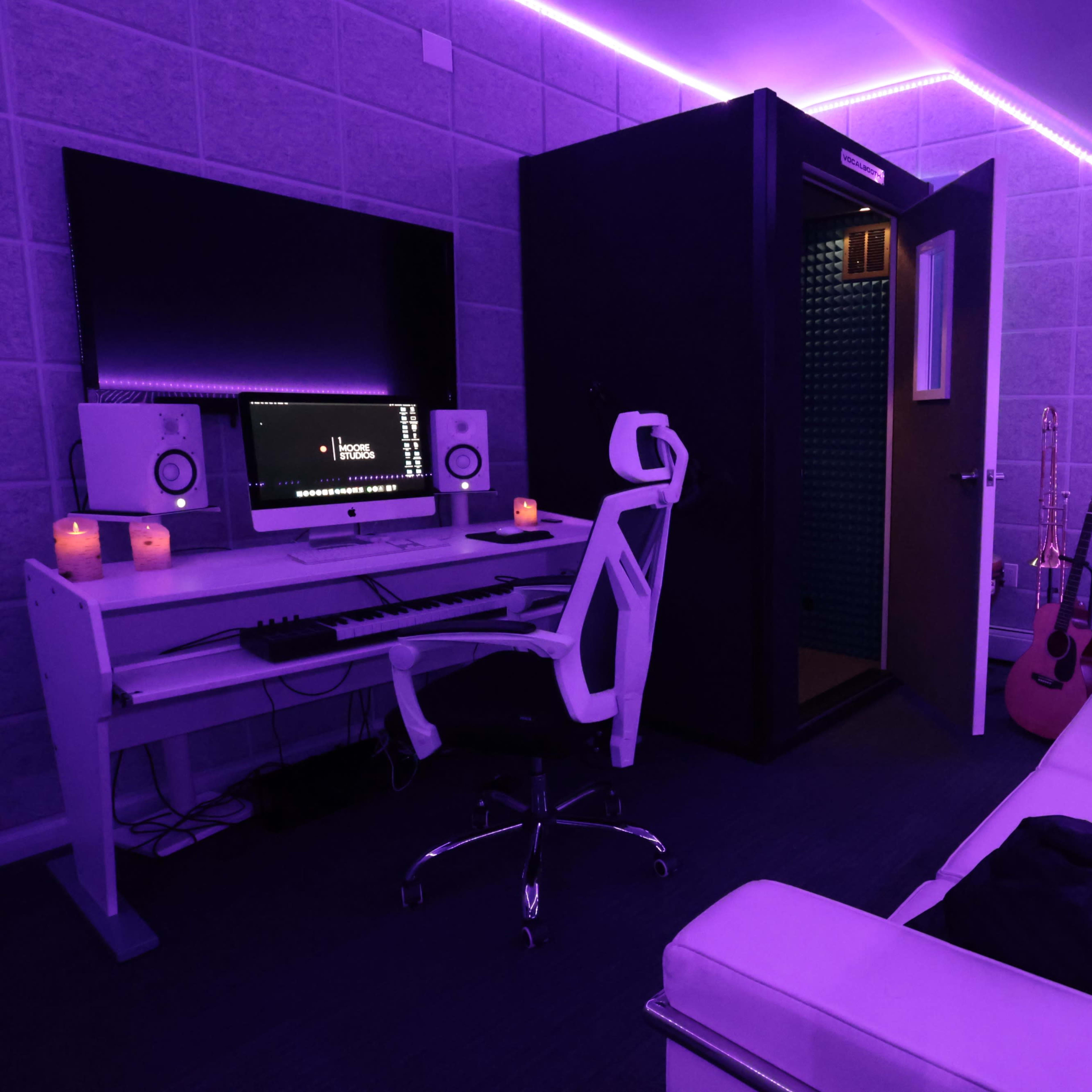 A music production room features a computer setup, soundproof booth, and cozy seating illuminated by purple lighting.