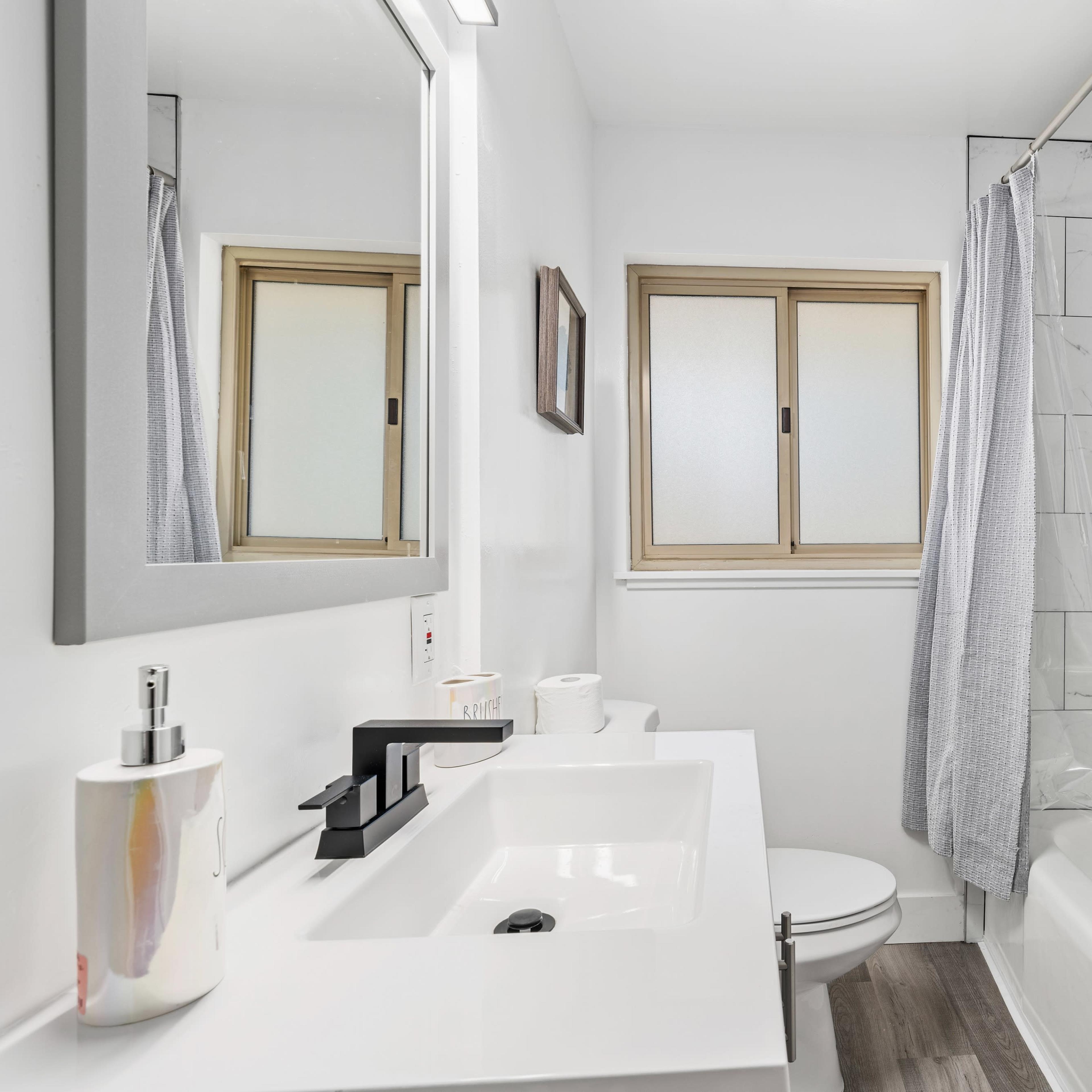 A modern bathroom with a rectangular white sink, a bathtub with a gray shower curtain, and a window above the tub.