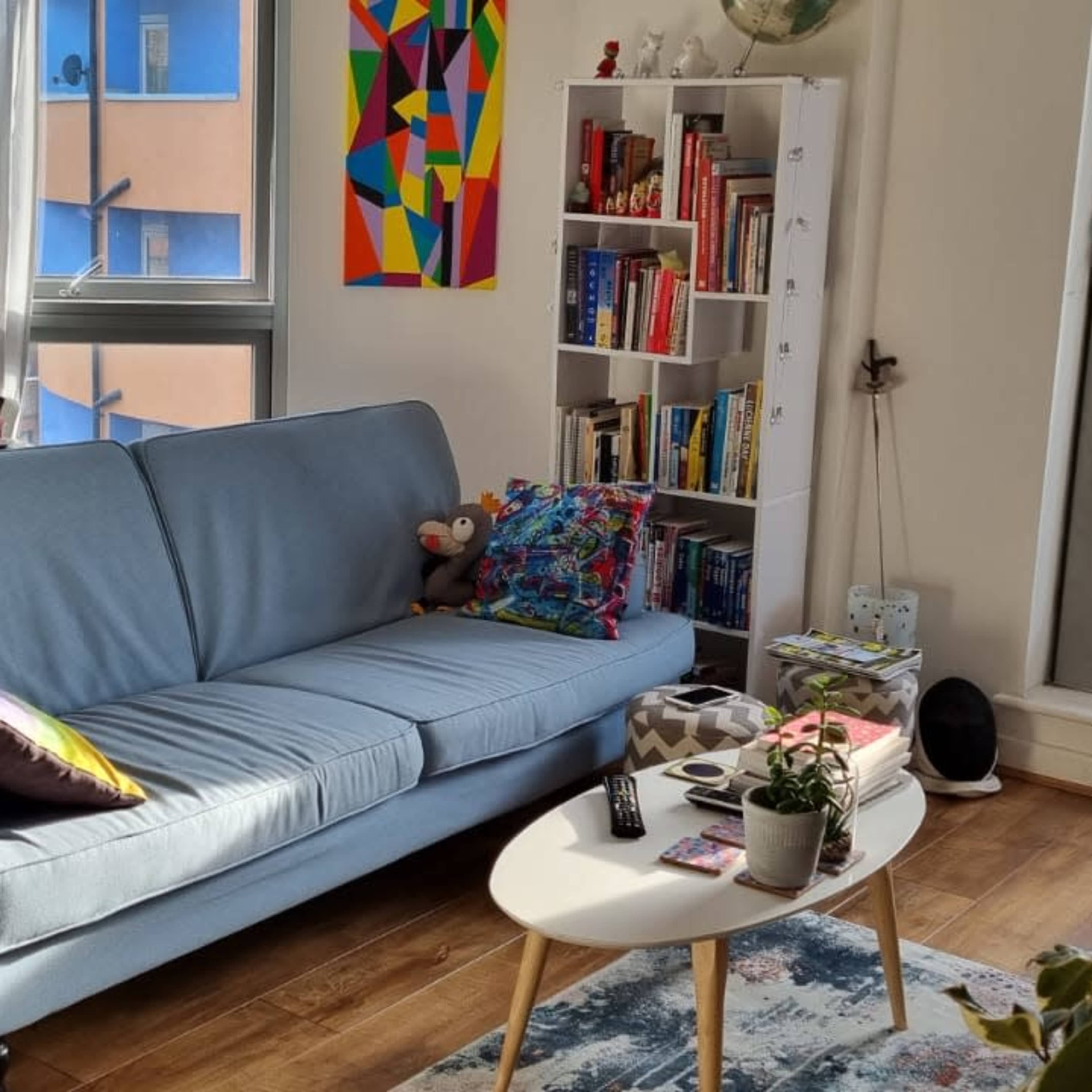 A bright living room with a light blue sofa, a dining table set for a meal, colorful wall art, and a bookshelf filled with books.