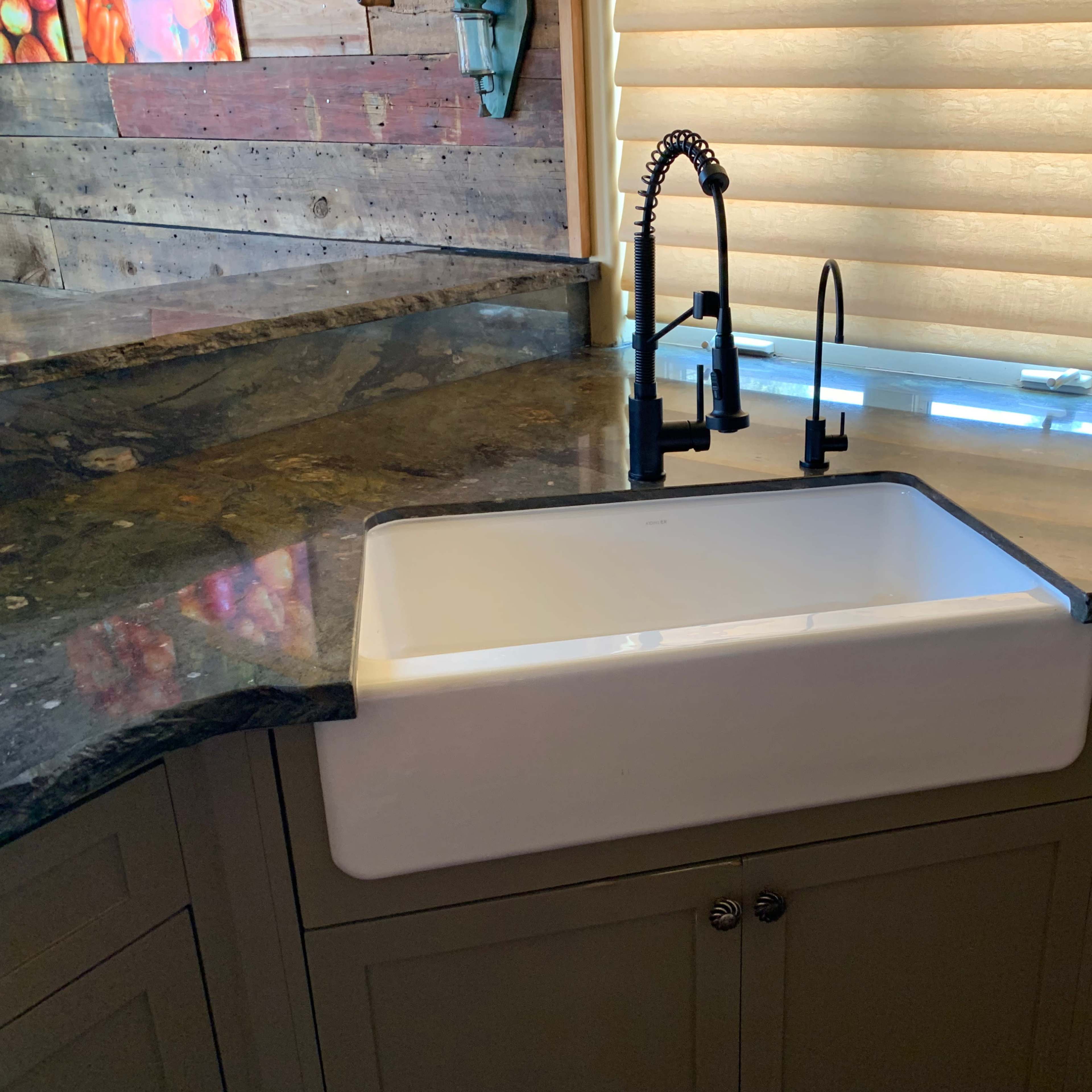 A farmhouse-style white sink is positioned within a kitchen countertop made of dark granite, featuring a black faucet and pull-down spray head.