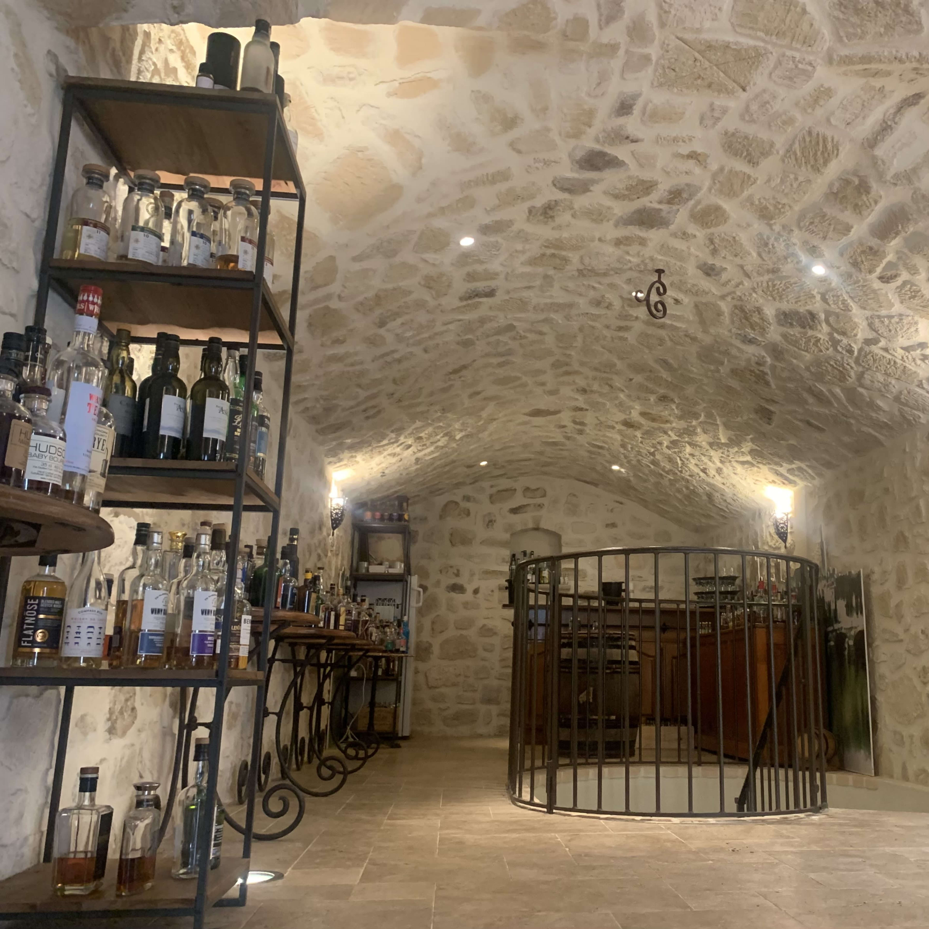 A stone-walled cellar with shelves of liquor bottles, wooden barrels, and a circular bar area in the center.