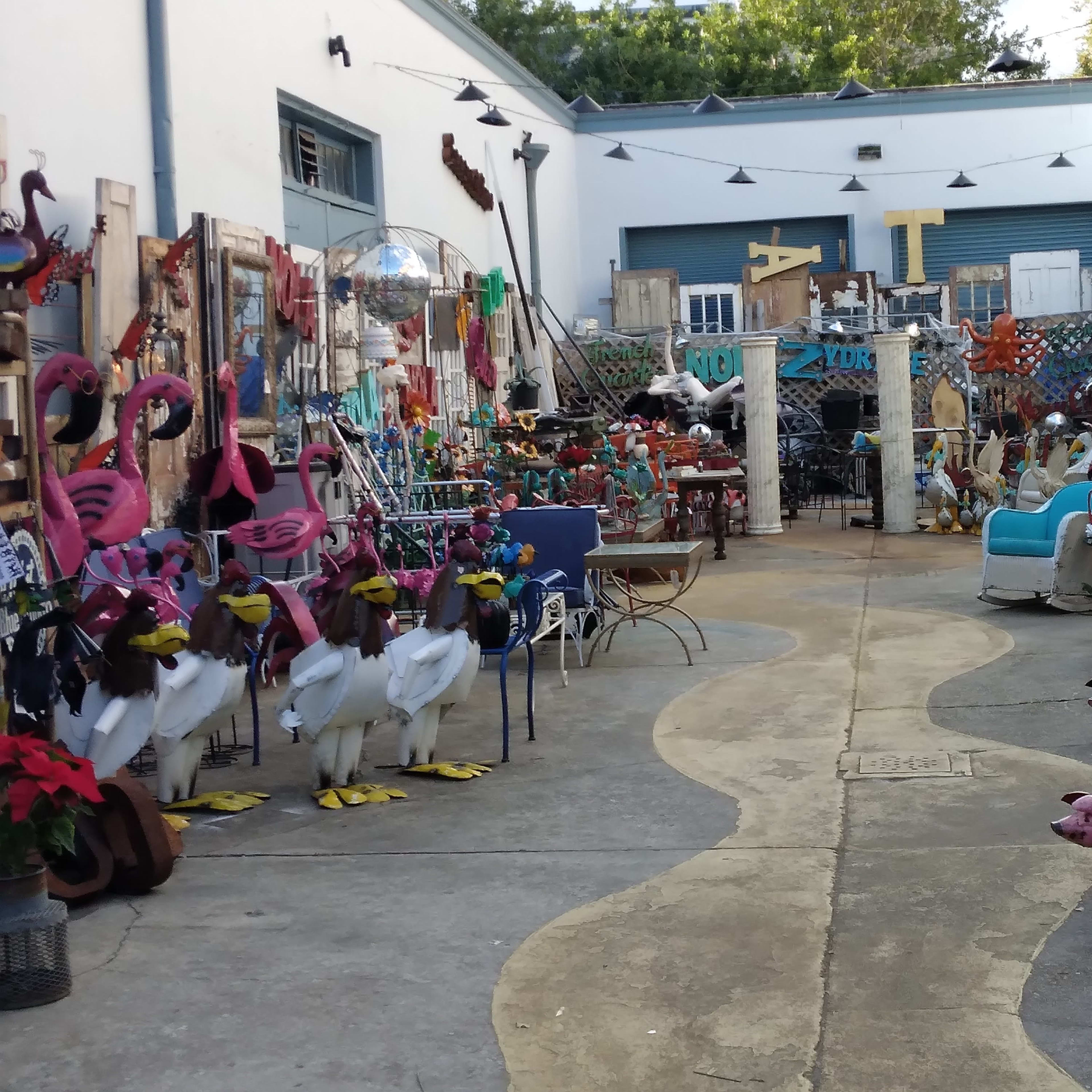 The scene features a colorful courtyard filled with various decorative items, including metal sculptures of flamingos, cows, pigs, and an assortment of furniture and art pieces.