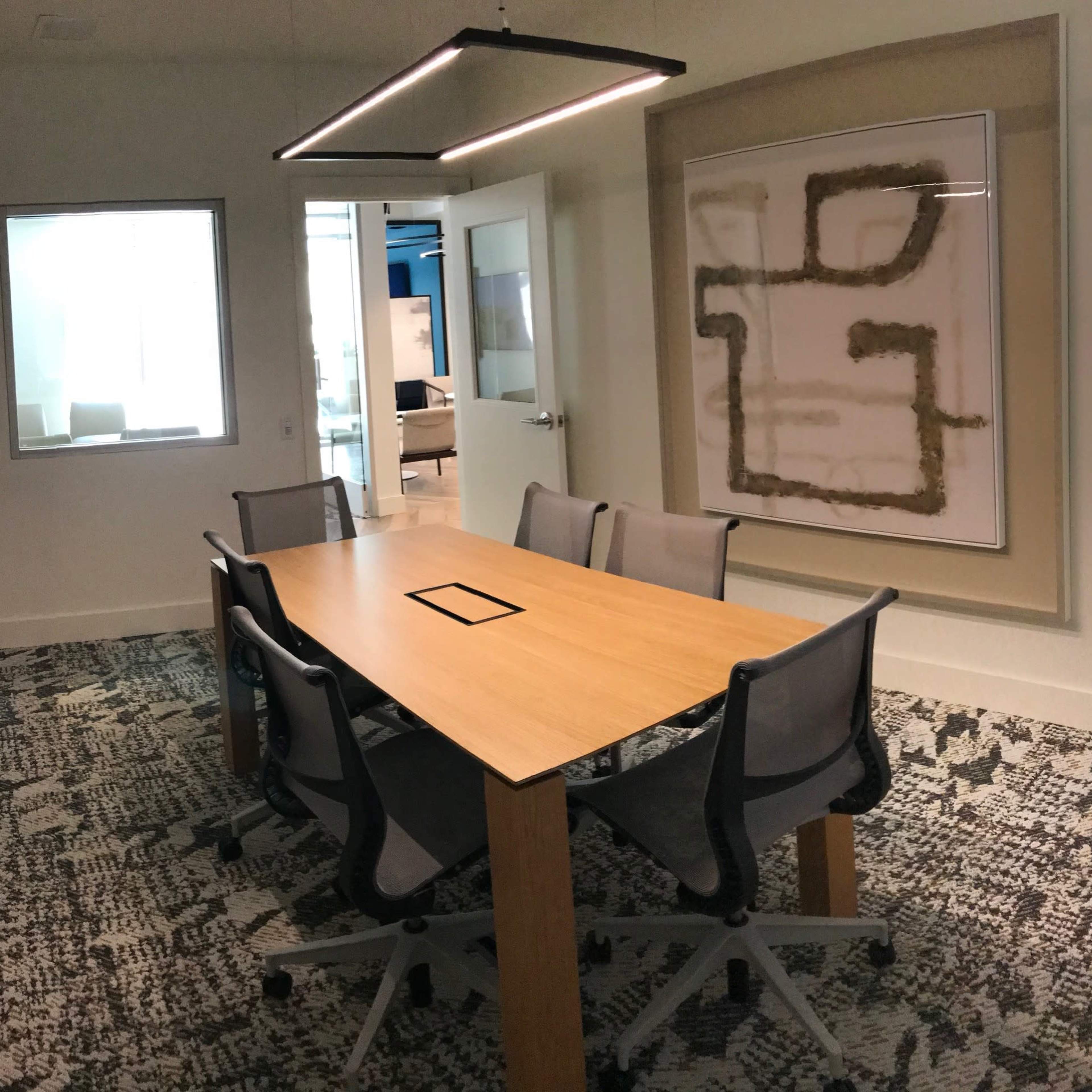 A conference room features a large wooden table surrounded by gray chairs, with a contemporary artwork on the wall and a window letting in natural light.