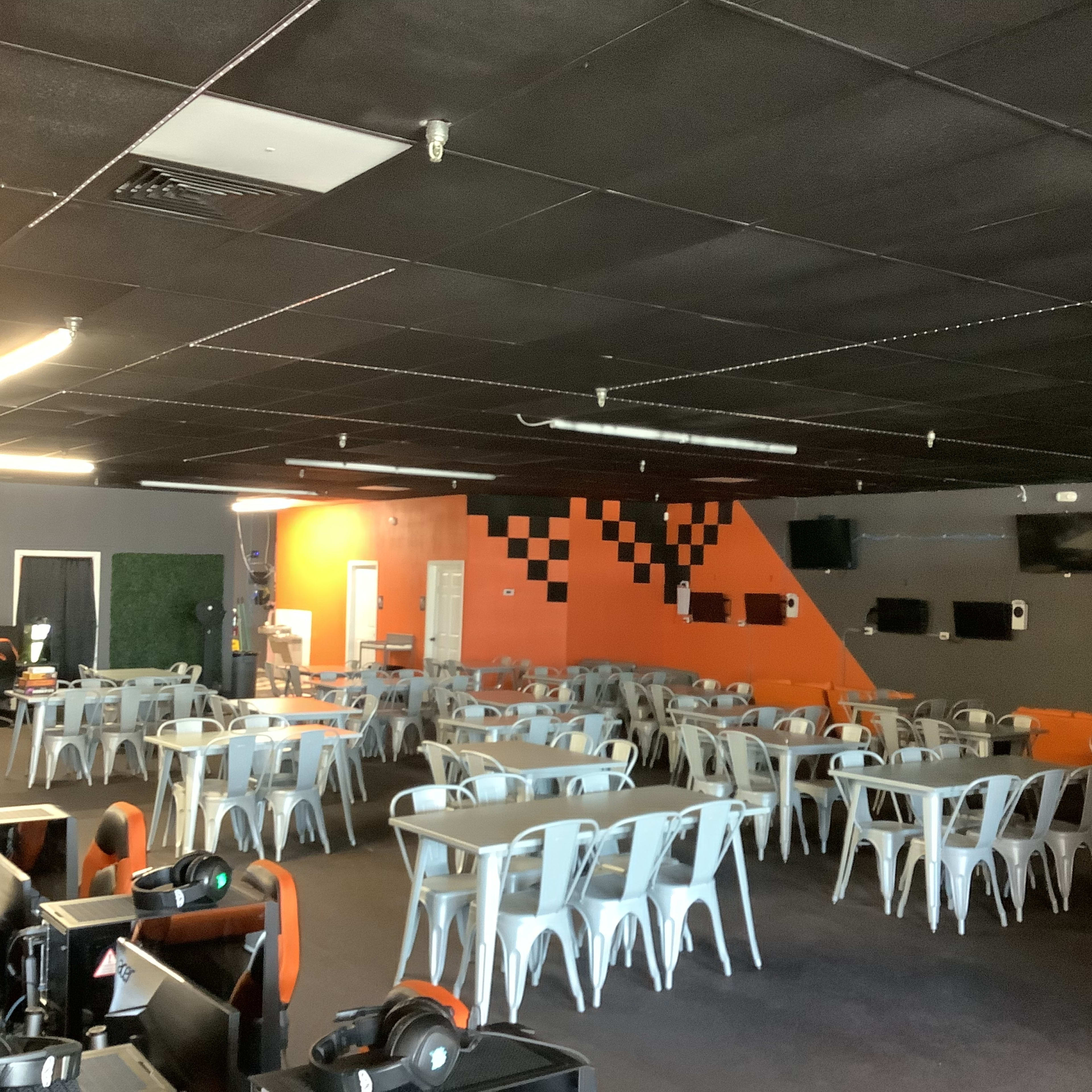 A spacious gaming room with multiple rows of metallic chairs and tables, orange and gray walls, and several screens mounted on the walls.