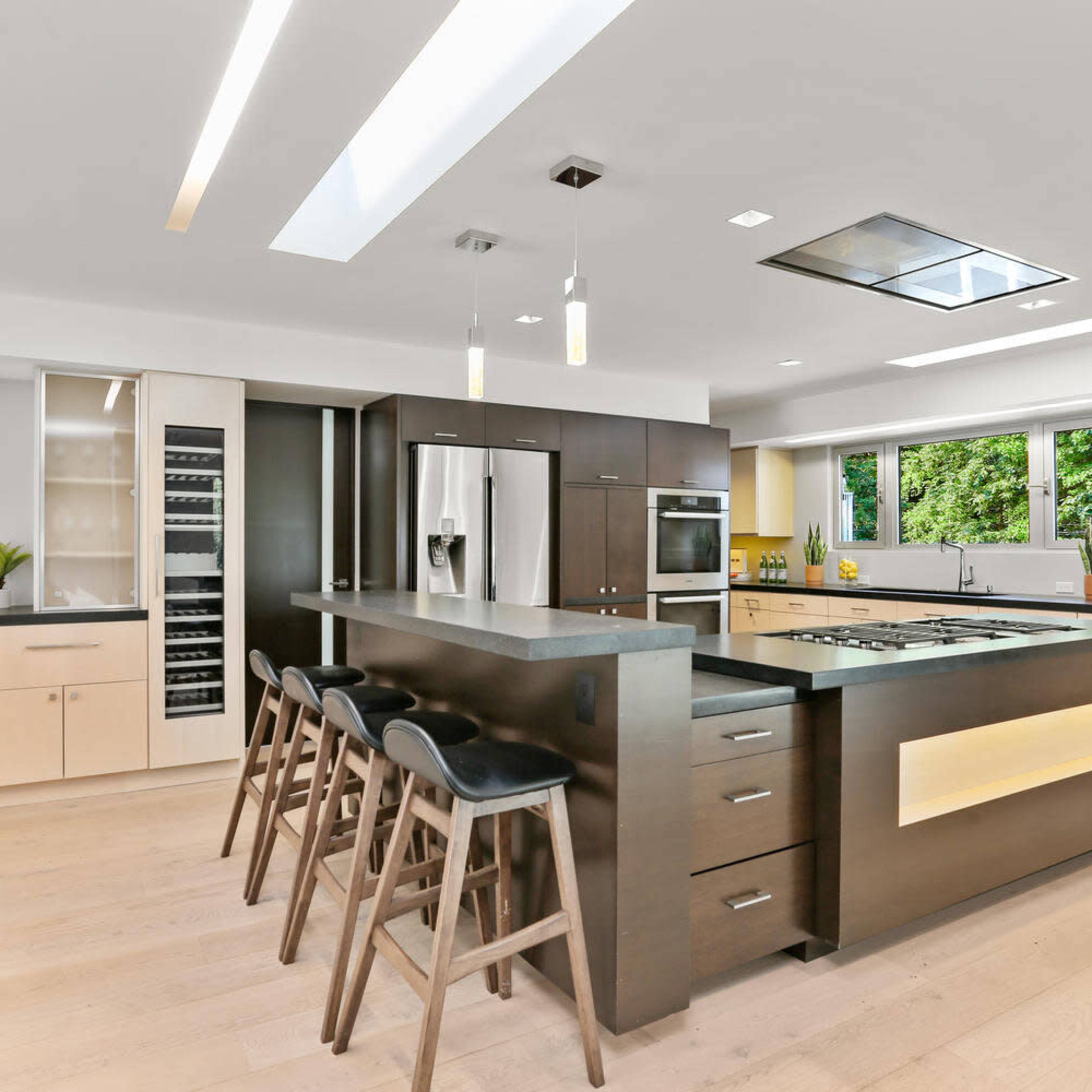 A modern kitchen features a central island with bar stools, dark cabinetry, and large windows allowing natural light.