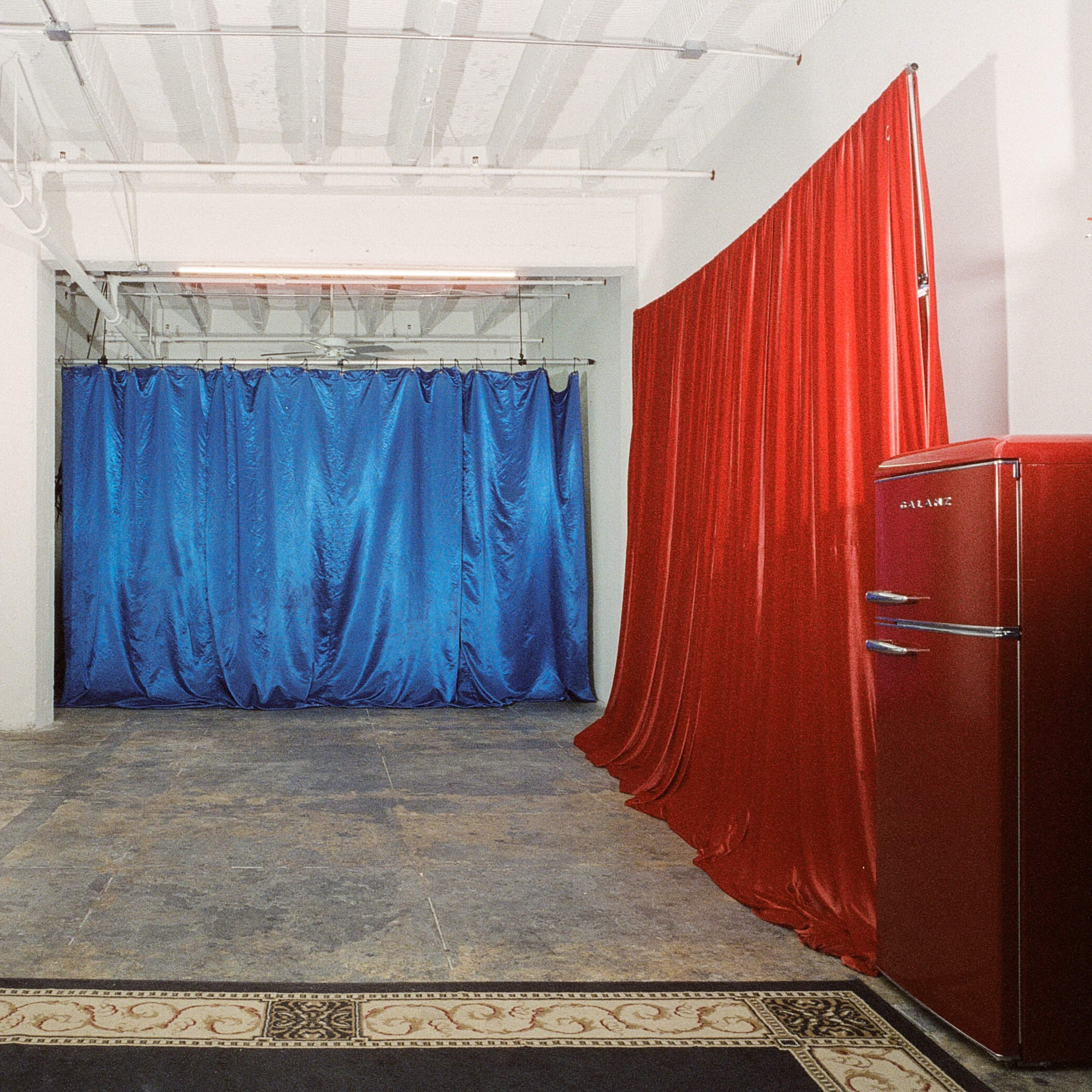 The scene shows a room with a concrete floor, draped red and blue curtains on the walls, and a vintage red refrigerator positioned in the corner.