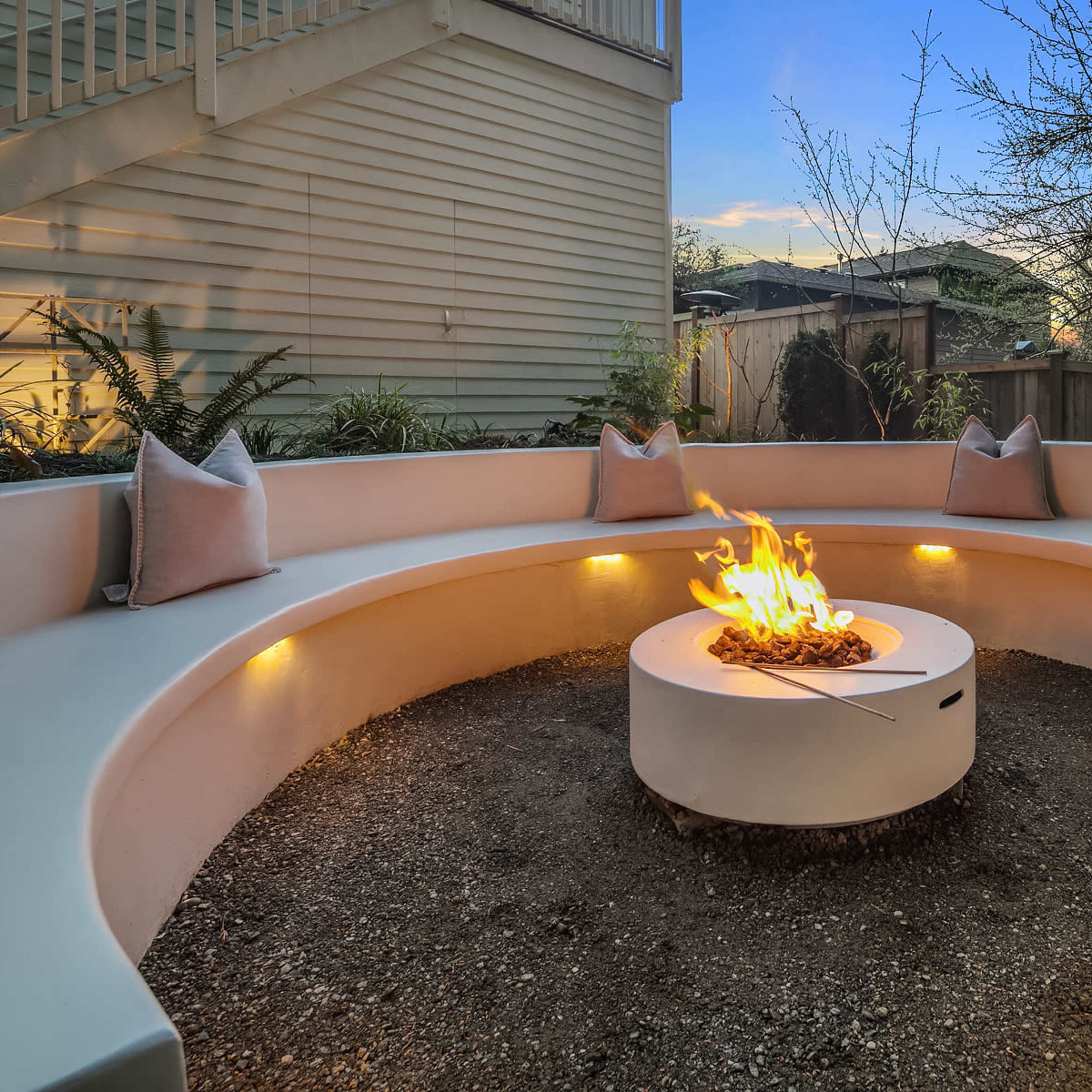A circular outdoor seating area with a central fire pit, surrounded by cushions and illuminated by soft lighting.