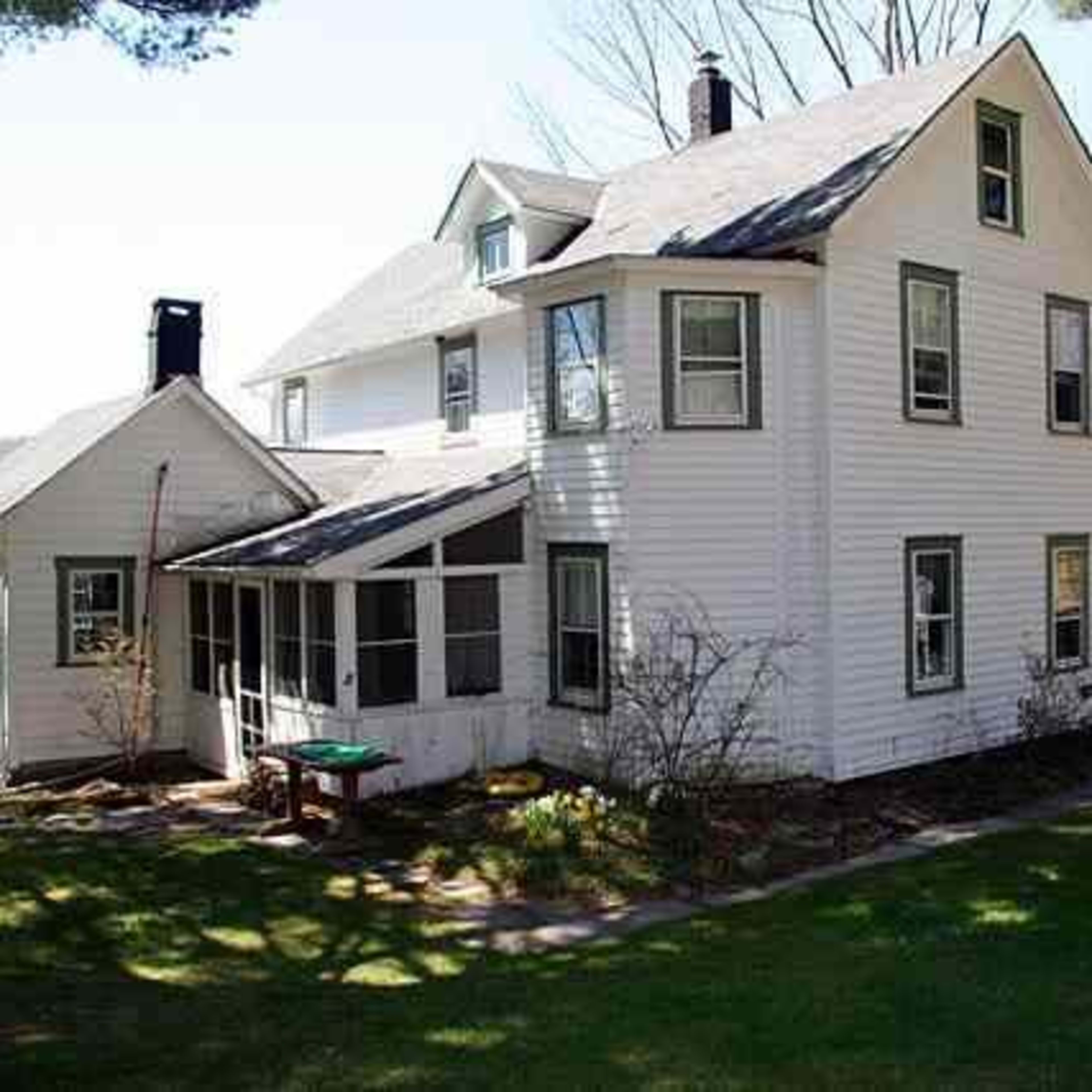 A two-story white house with a large porch and several windows stands in a grassy area, surrounded by trees and a scenic background.