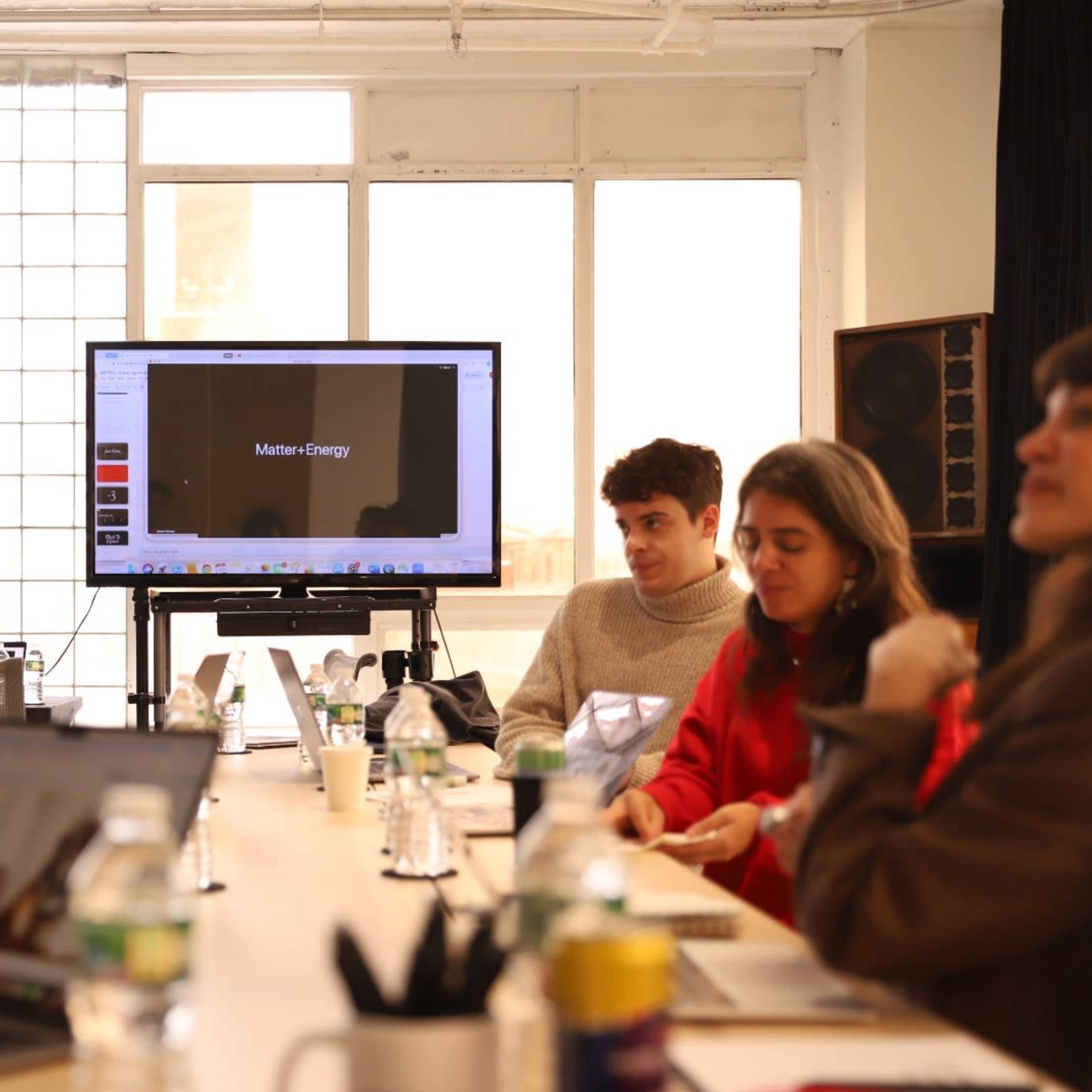 A group of five people is seated around a table during a meeting, with a presentation screen displaying a title in the background.