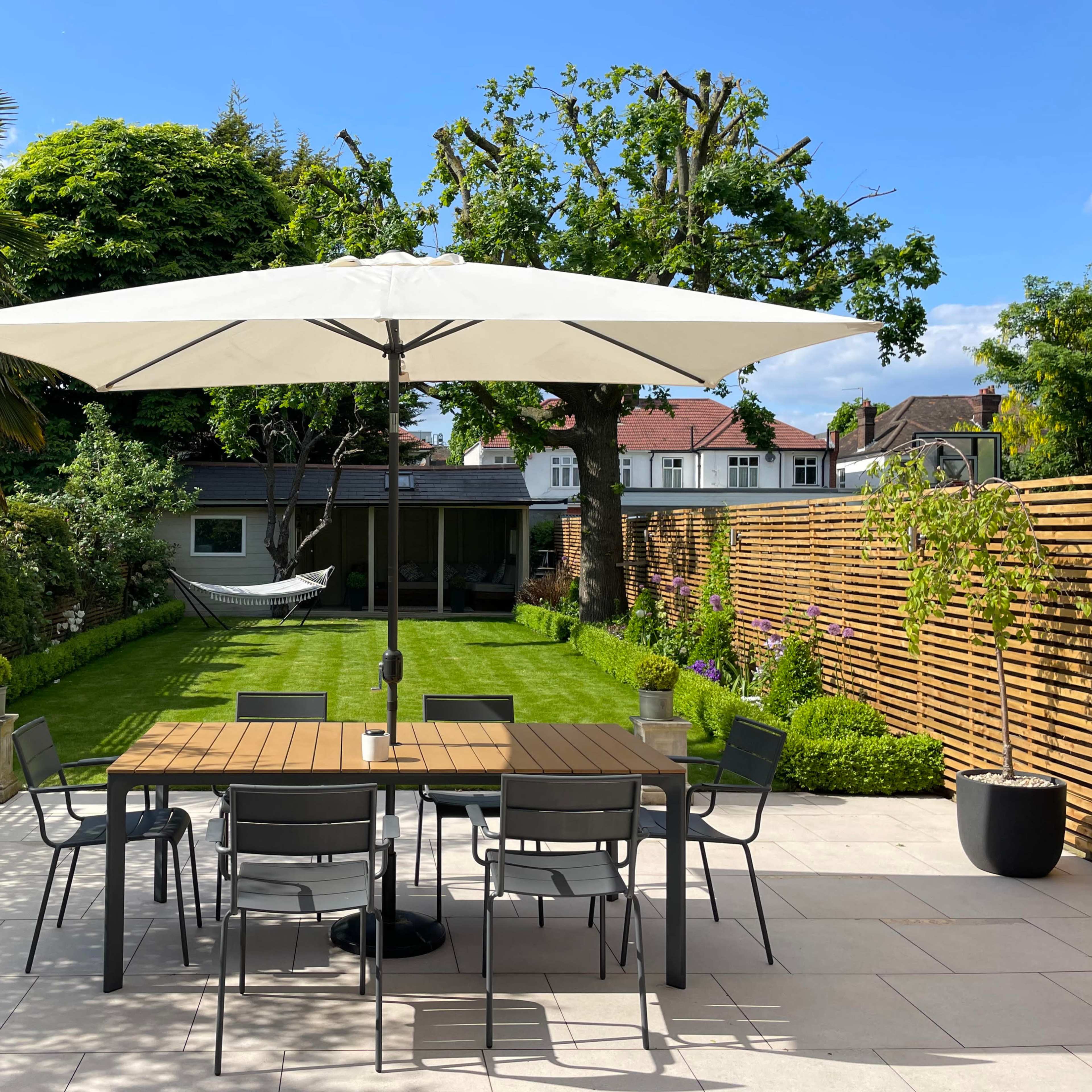 A spacious garden features a large table with chairs under a white umbrella, surrounded by neatly trimmed grass and trees.