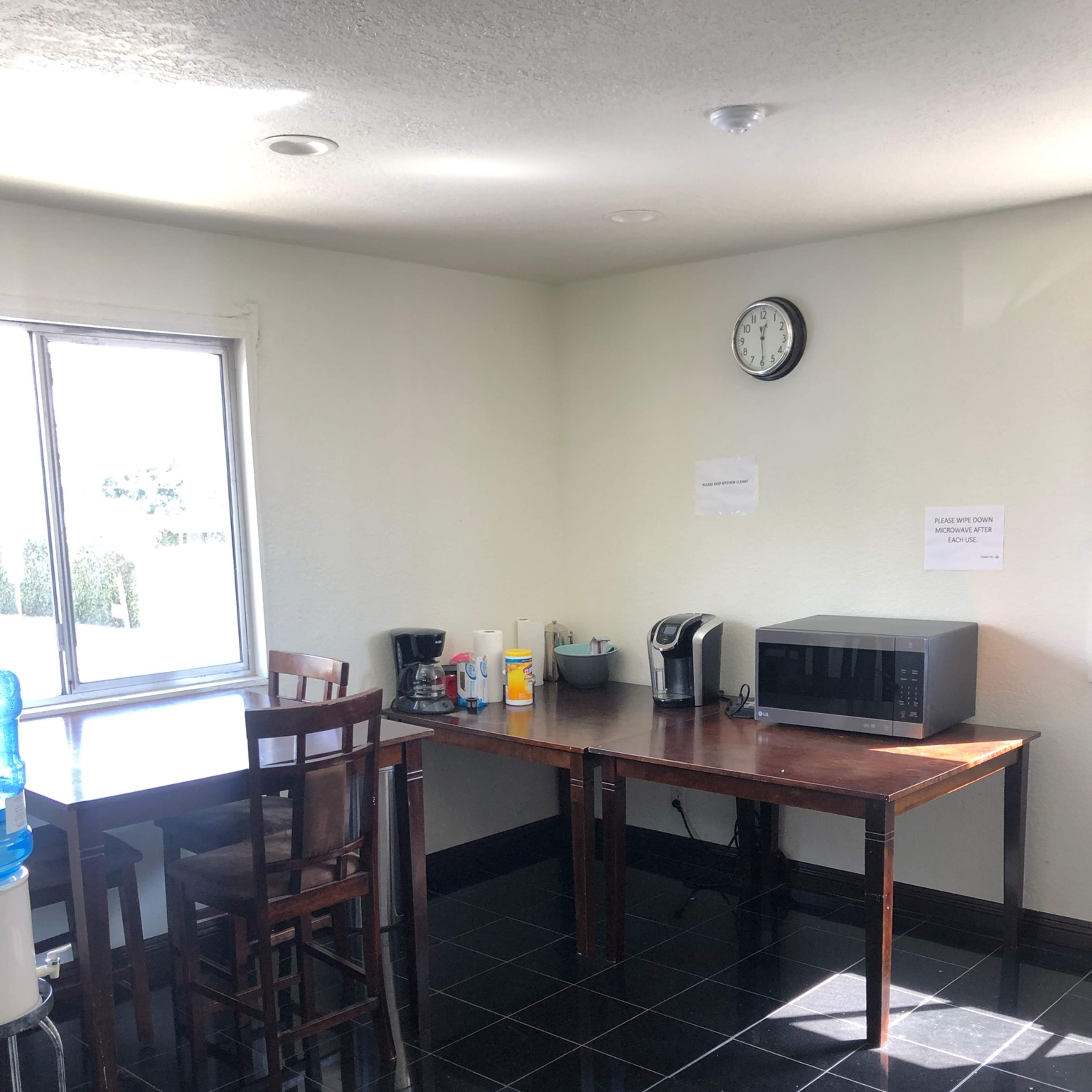 A simple kitchen area with two tables, a water cooler, a coffee maker, a microwave, and a clock on the wall.