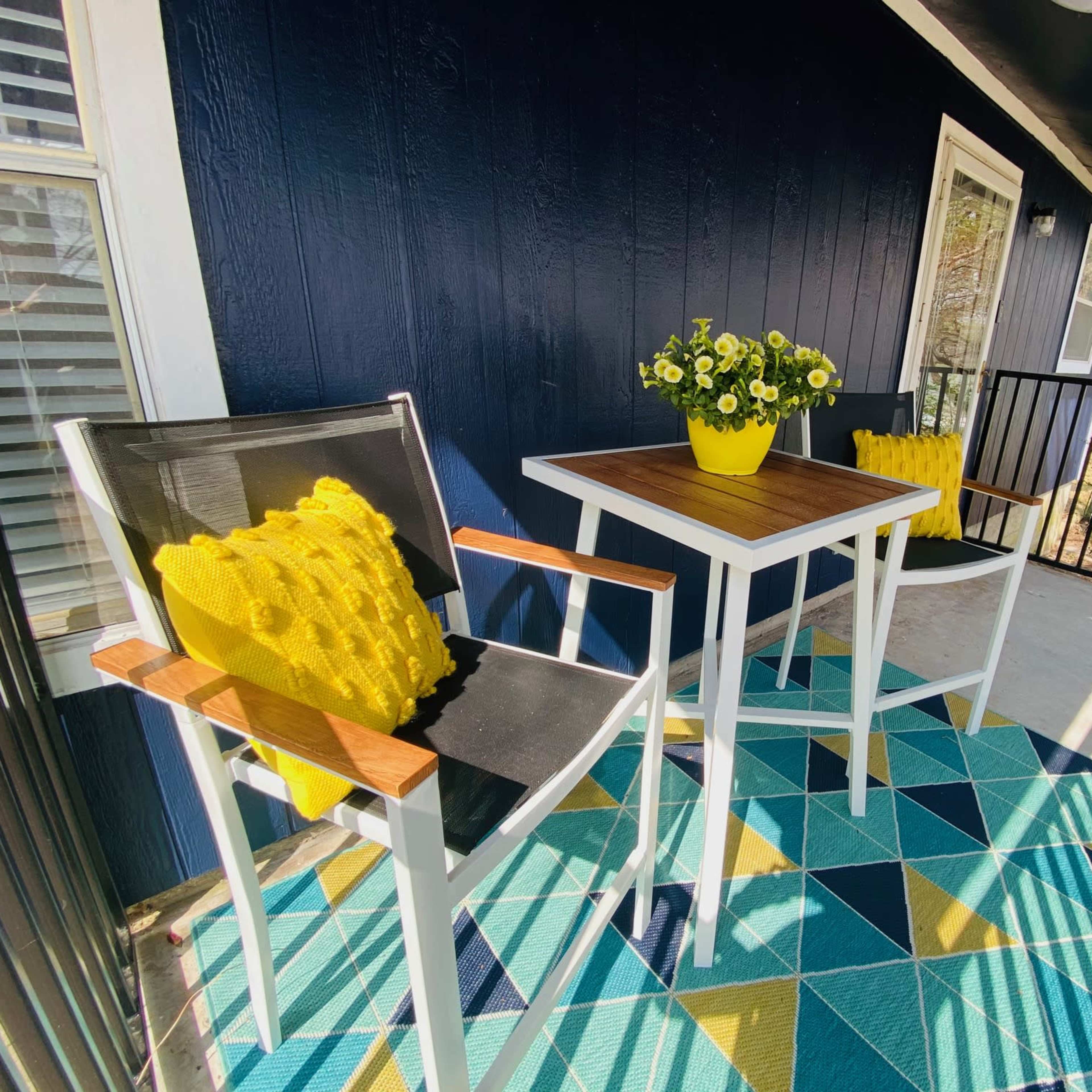 A small balcony features two chairs and a table, adorned with a yellow flower pot and a yellow cushion on one chair, set atop a geometric-patterned rug.