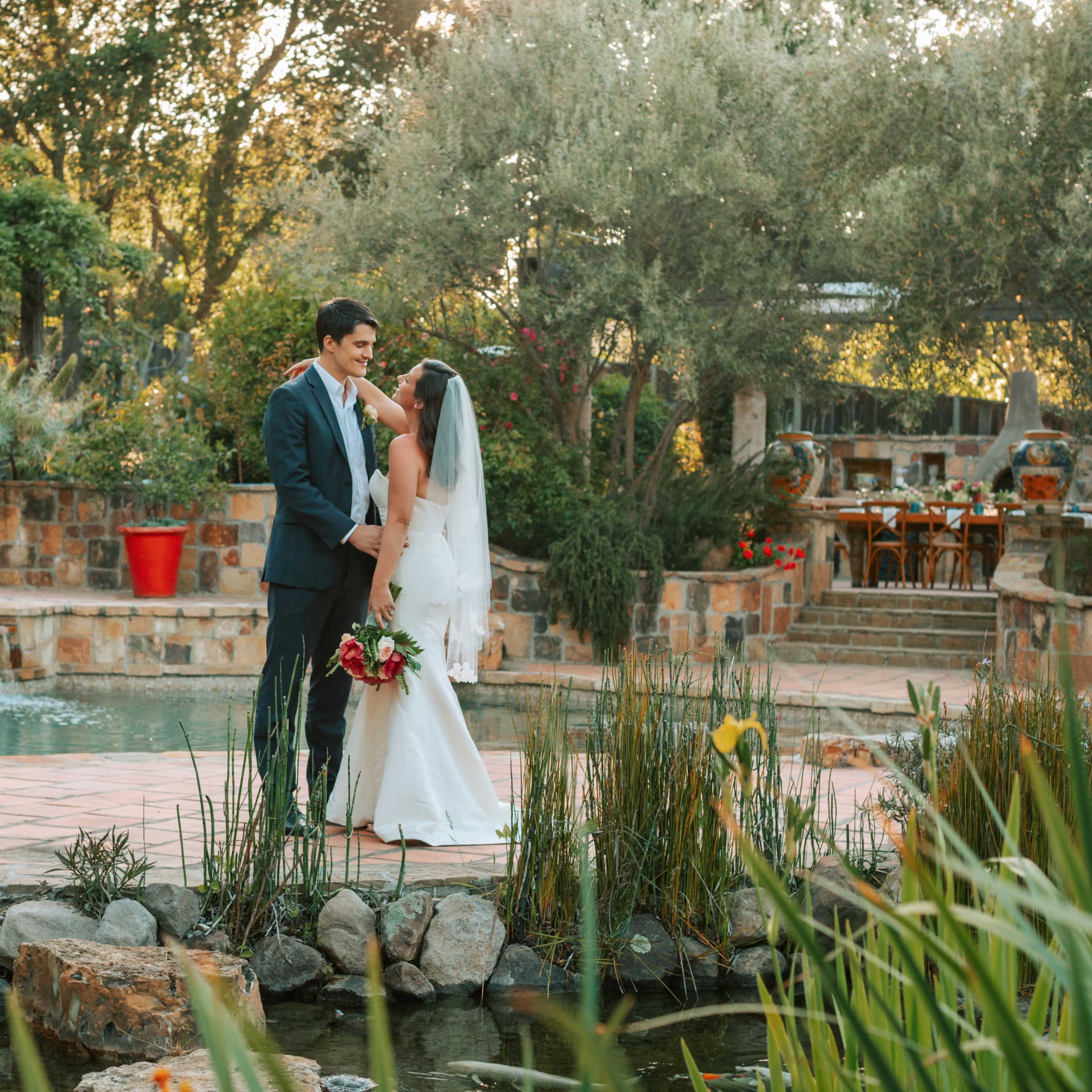 A bride and groom stand close together on a stone path beside a tranquil pond, surrounded by lush greenery and elegant wedding decor.