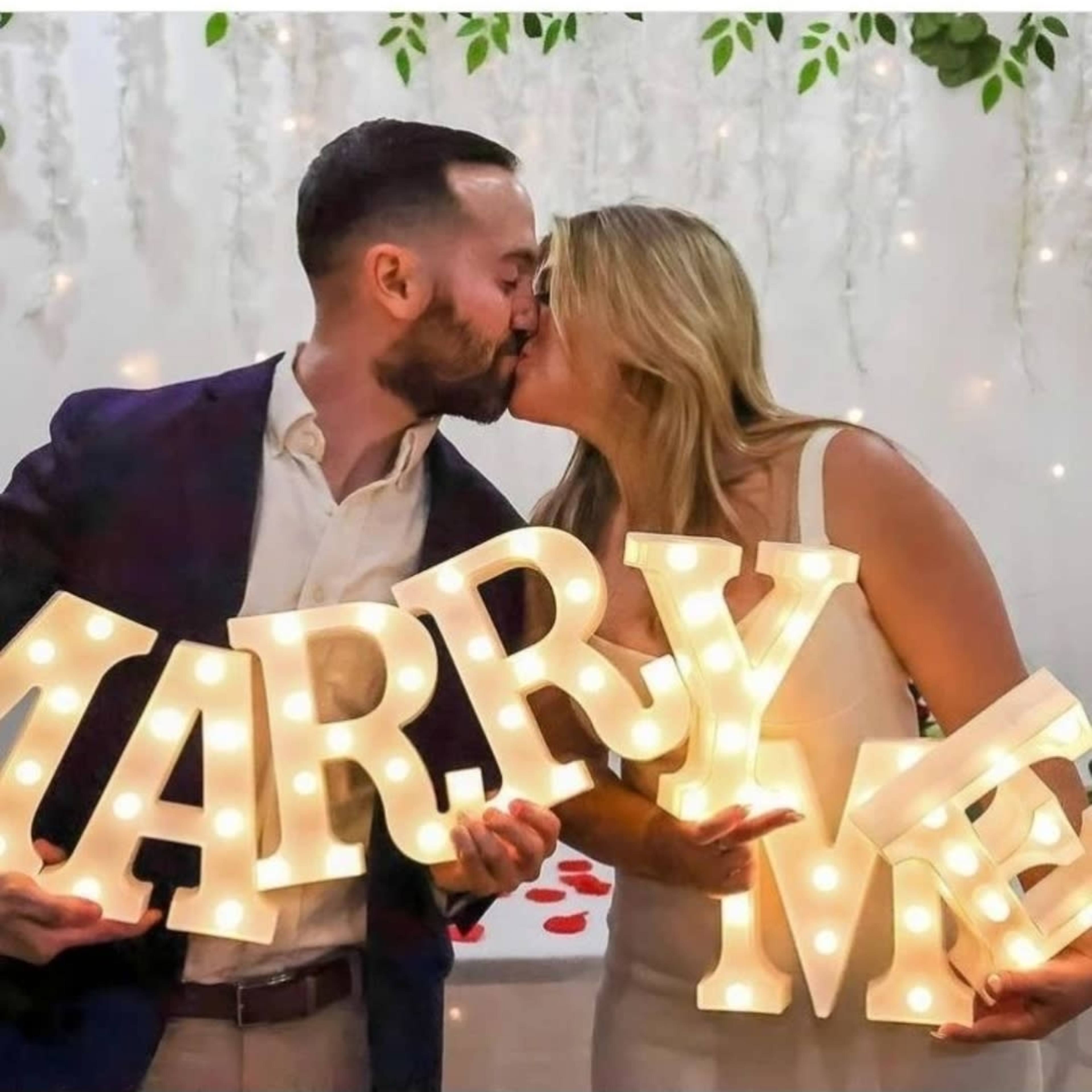 A couple is kissing while holding illuminated letters that spell "MARRY ME."