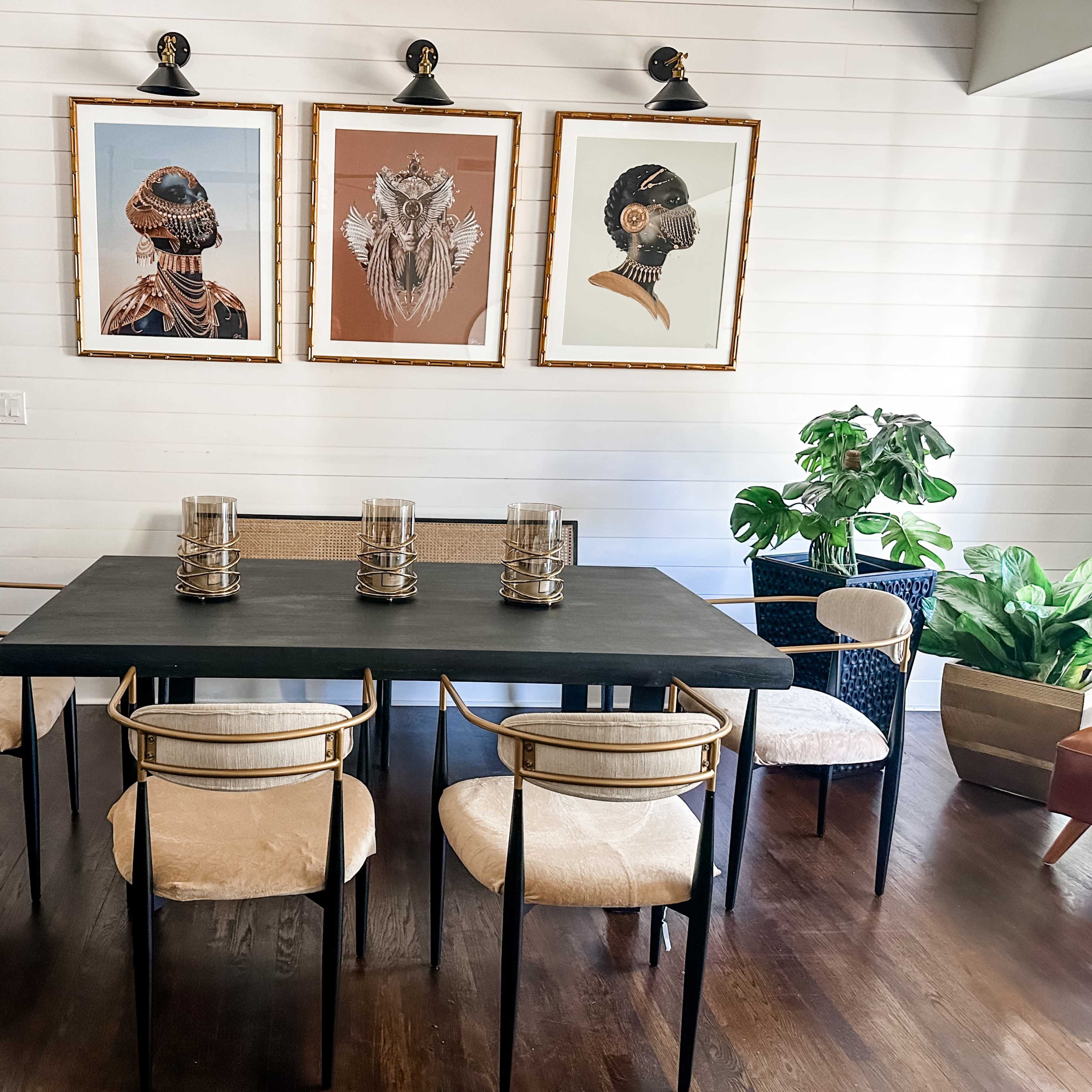 A dining area with a black table surrounded by six chairs, two of which have a light upholstery, and three framed artworks on the wall above.