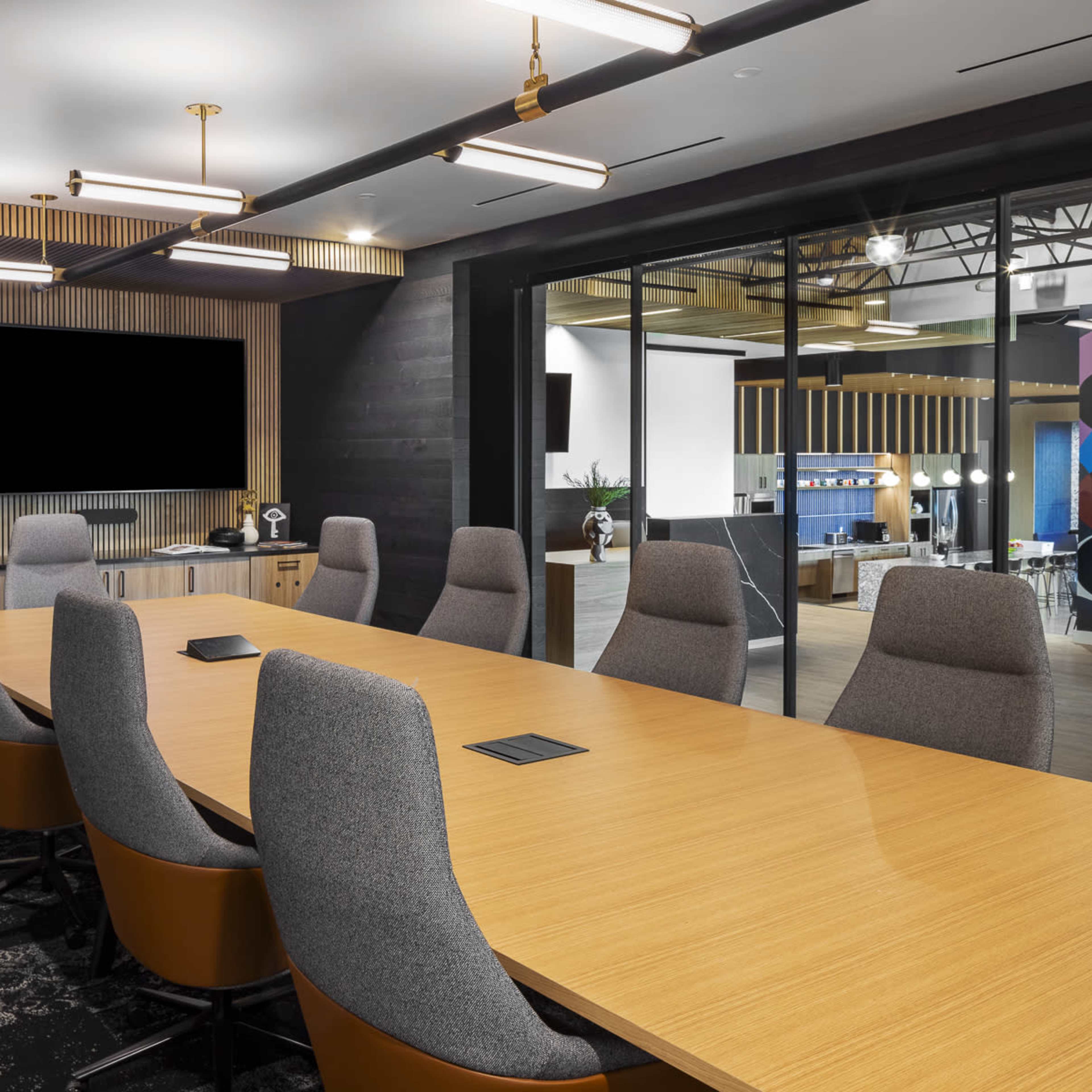 A modern conference room with a long wooden table surrounded by gray upholstered chairs, featuring two large blank screens on the wall.