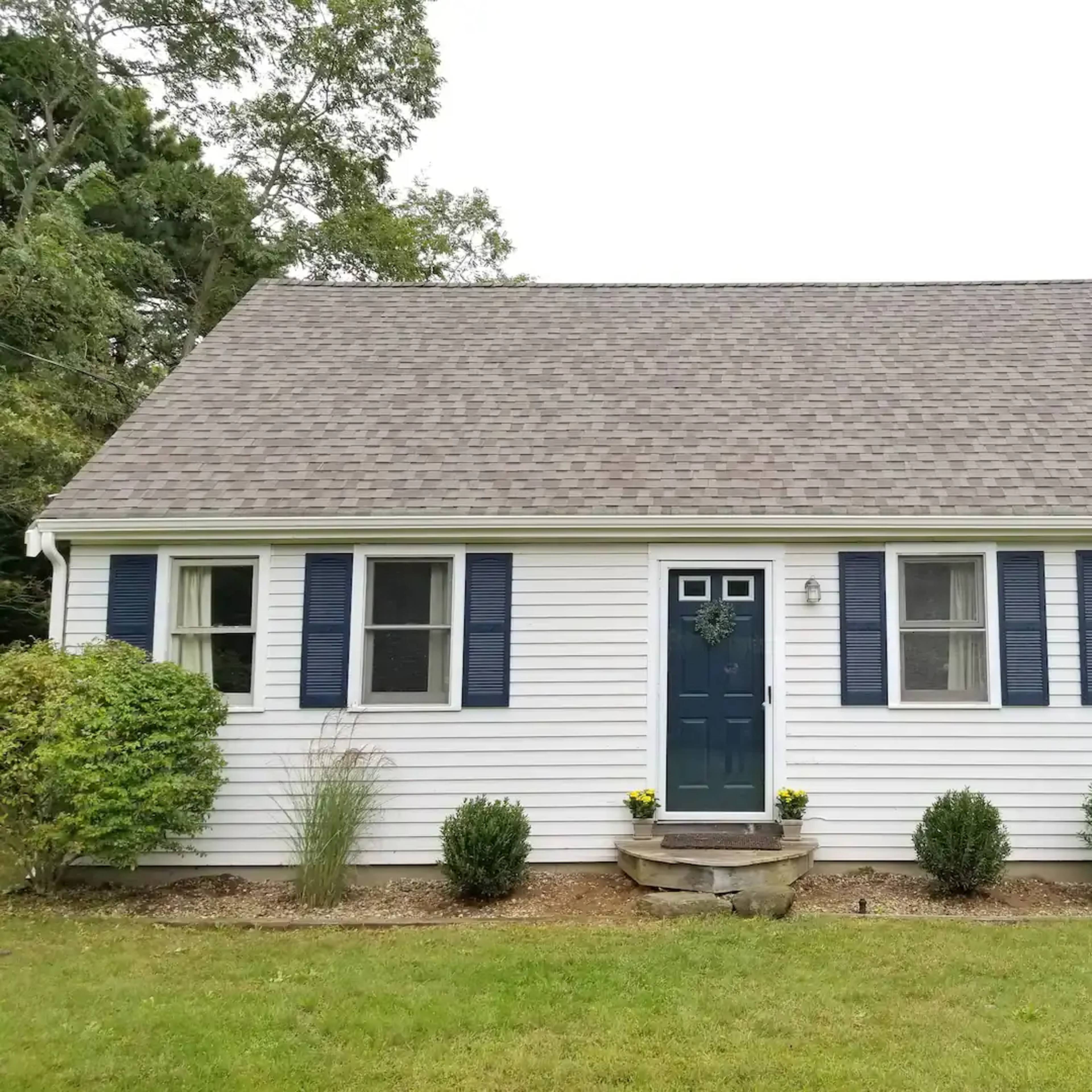 A single-story house features a blue front door, white siding, and dark blue shutters on the windows, surrounded by neatly trimmed shrubs.