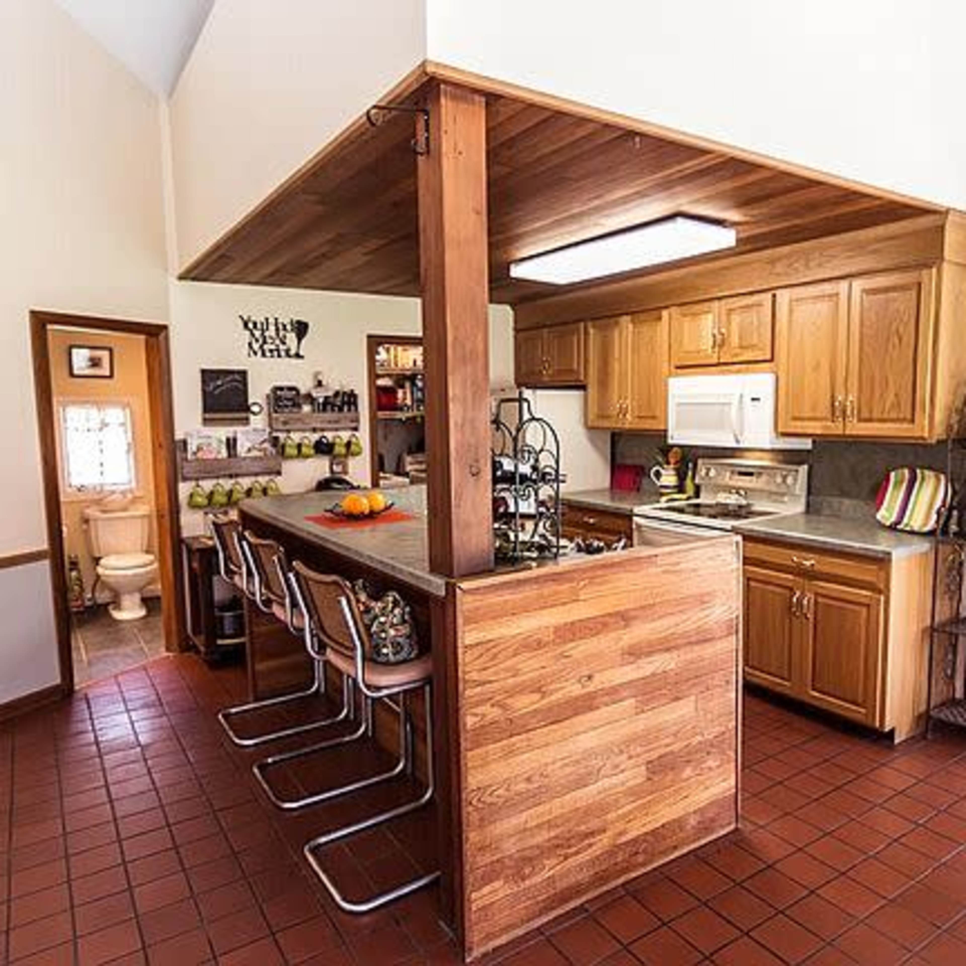 A kitchen with wooden cabinets, a central bar with stools, and a dining area in the background.