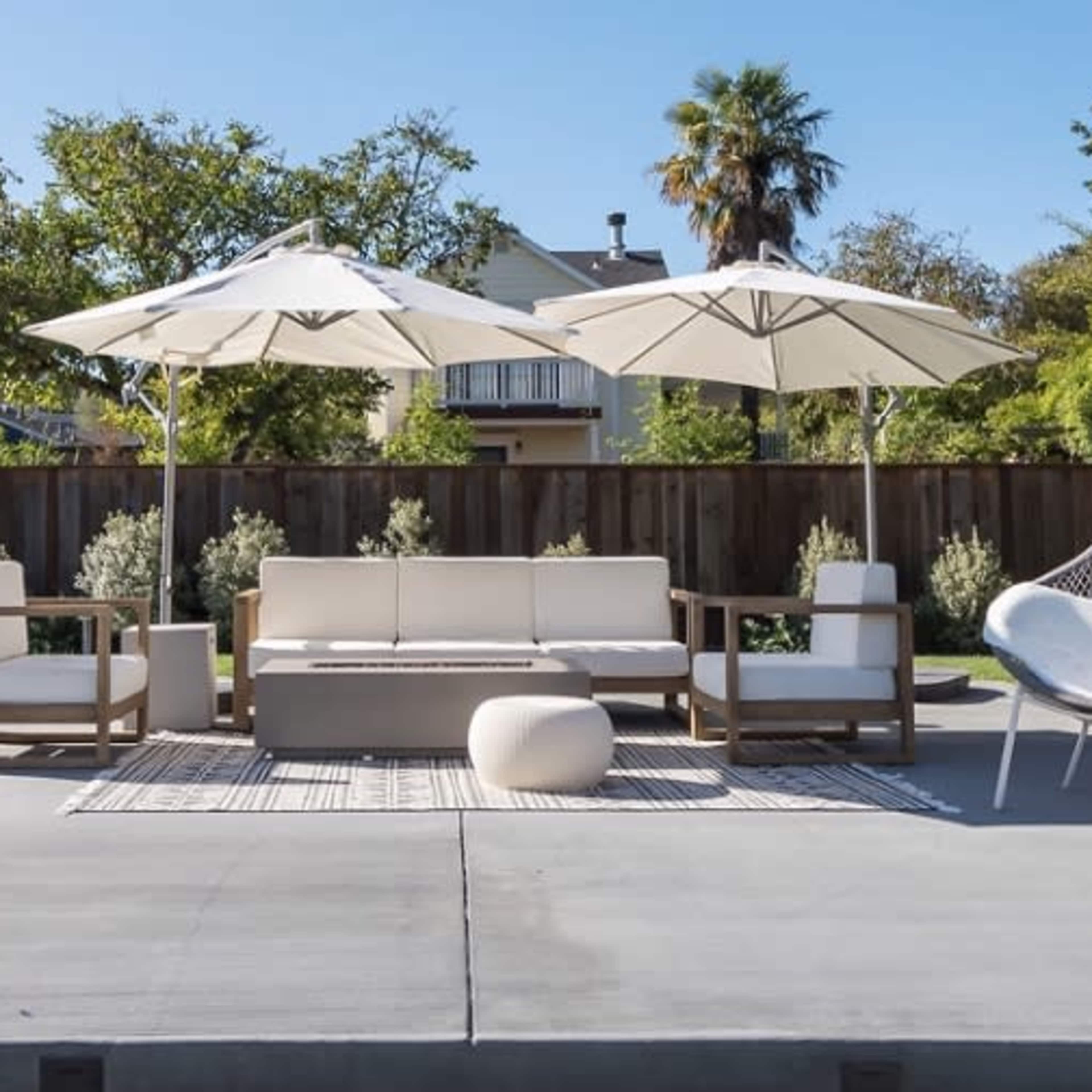 An outdoor seating area with a white couch, wooden chairs, and large umbrellas surrounded by greenery and a concrete patio.