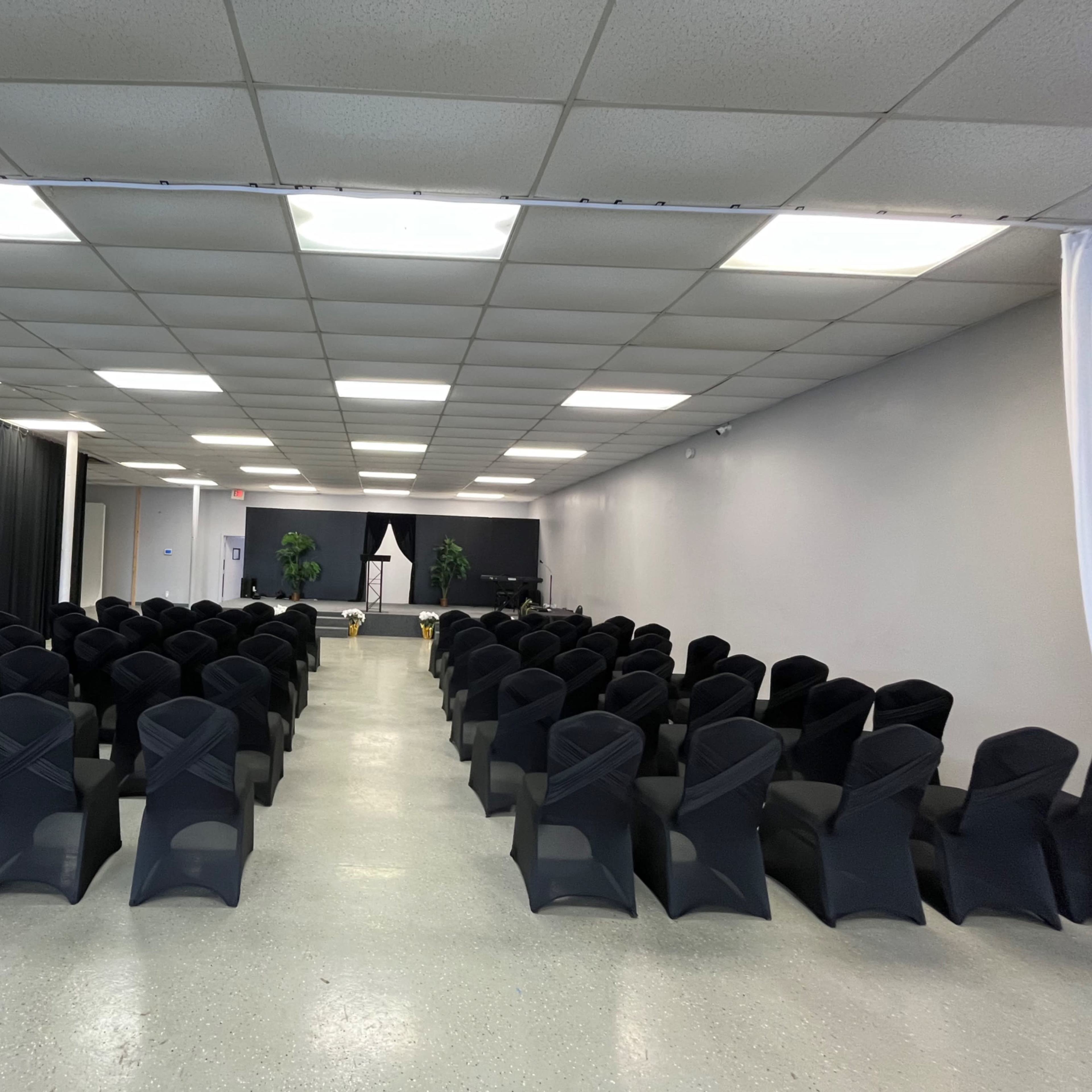 A spacious room with rows of black chair covers arranged neatly facing a stage area, flanked by potted plants.