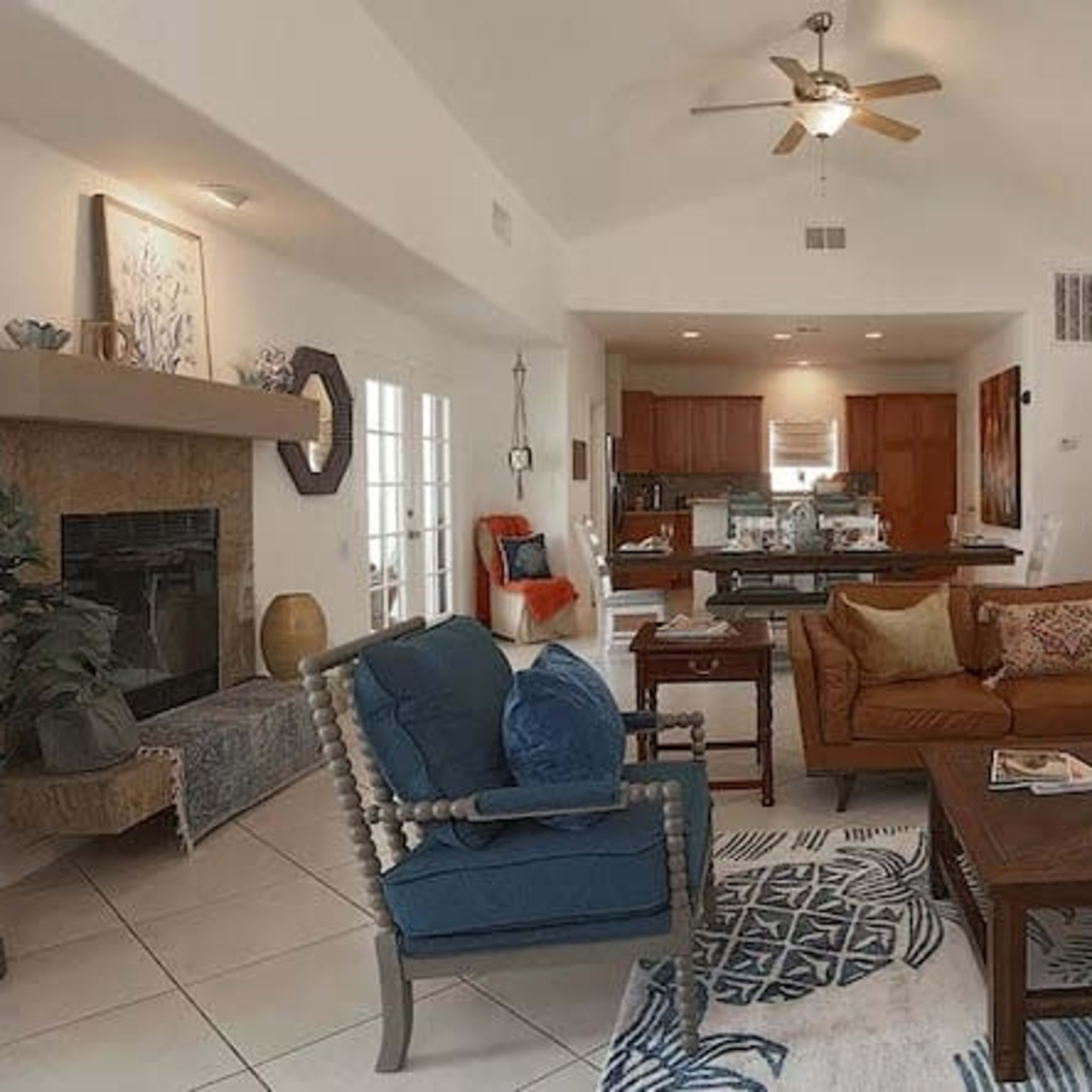 A living room features a brown leather sofa, blue armchairs, a wooden coffee table, and a kitchen in the background with light-colored cabinetry.
