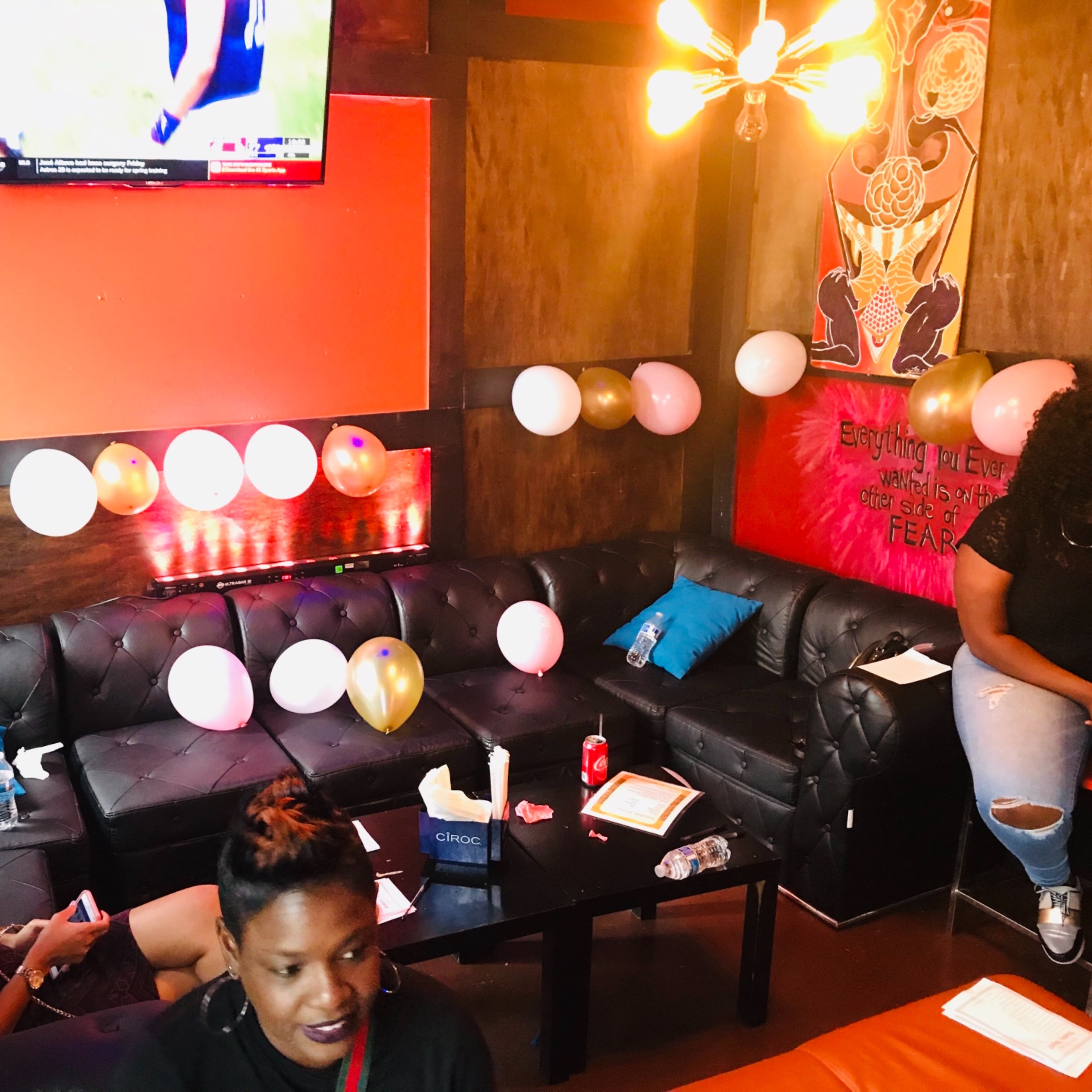 A small, decorated lounge area features black leather seating, colorful balloons, and a television displaying a sports event on the wall.