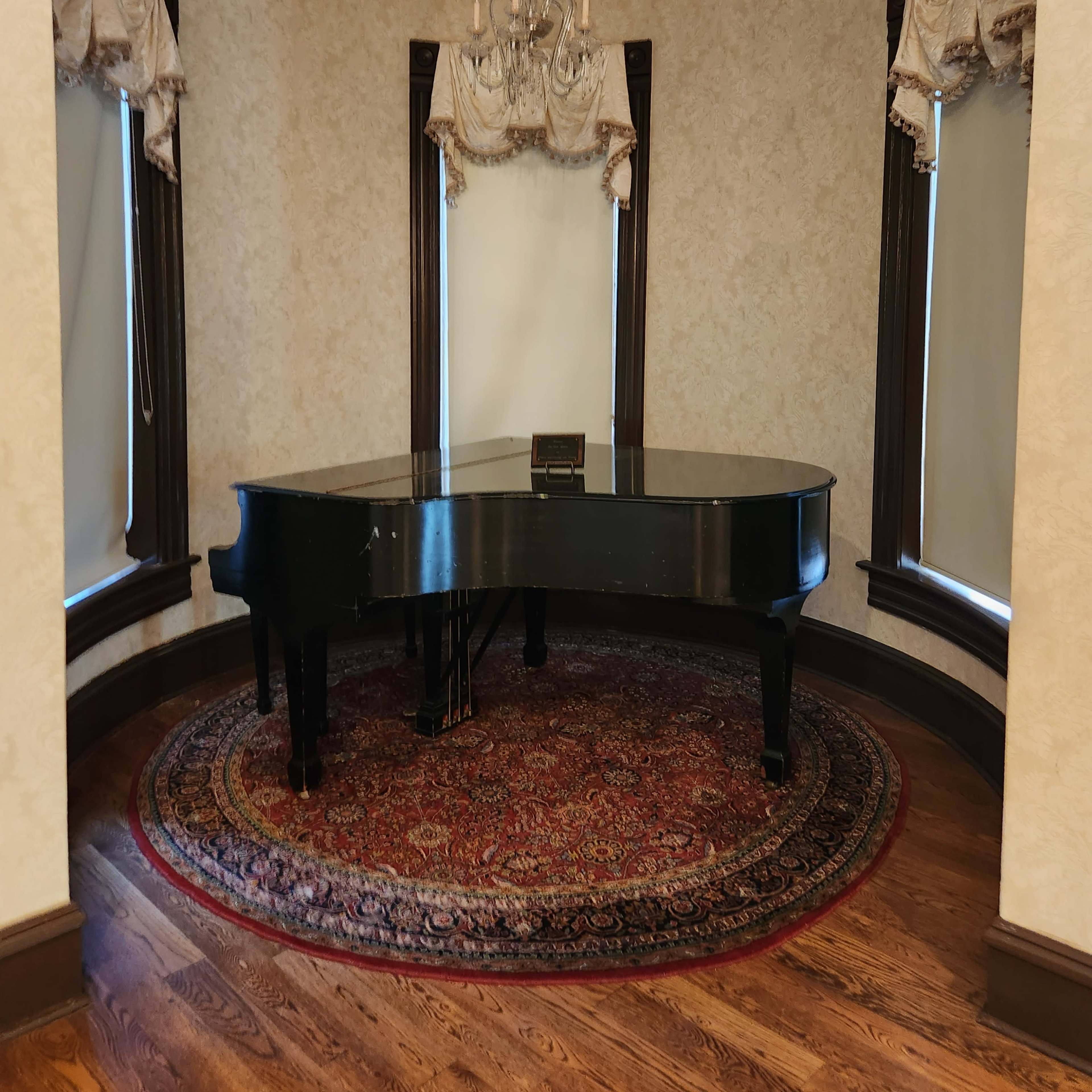 A black grand piano is situated in a corner of a room adorned with a patterned rug and large windows covered by drapes.