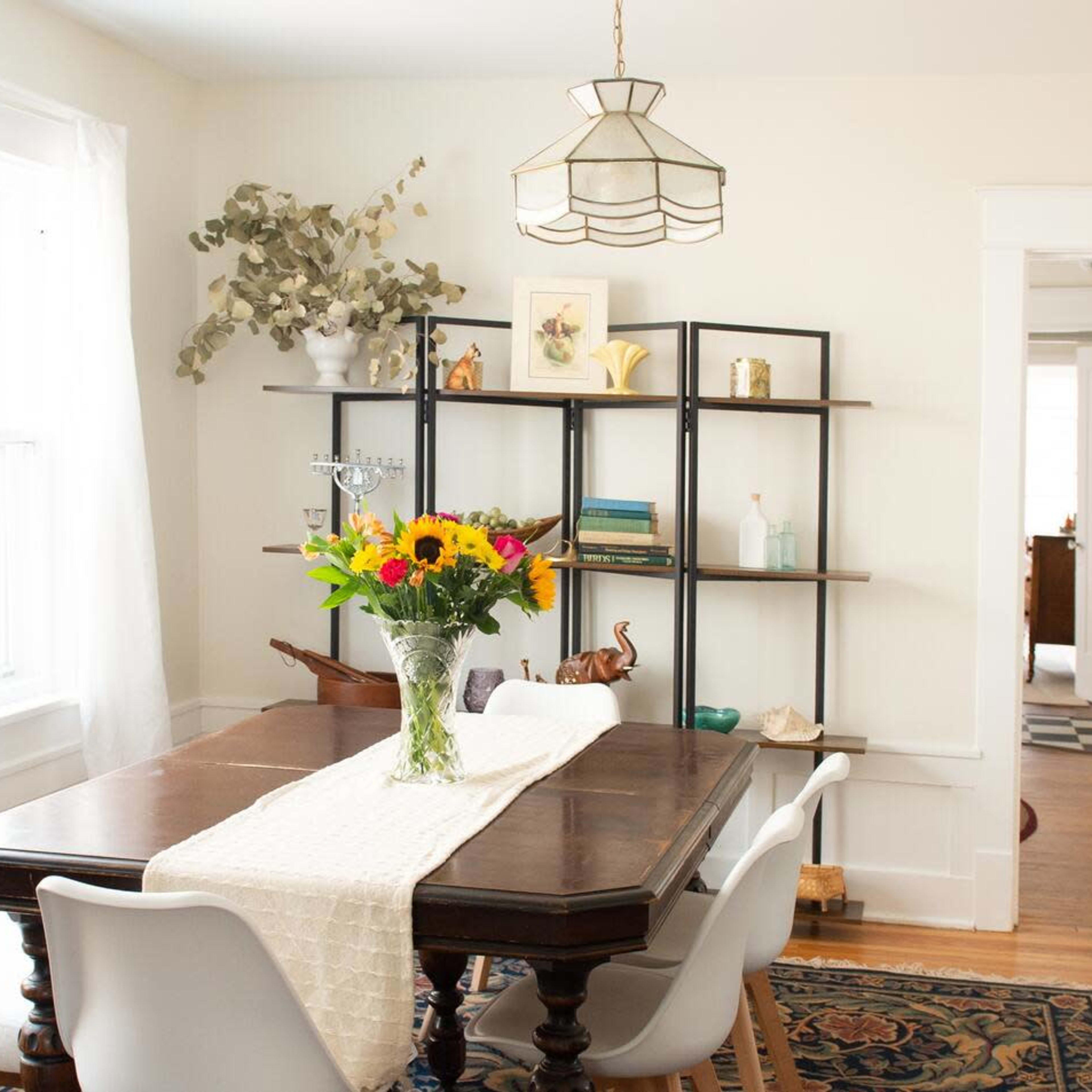 A dining area features a wooden table with a floral centerpiece, surrounded by white chairs, and a shelf displaying various decorative items, with a flag hanging in the background.