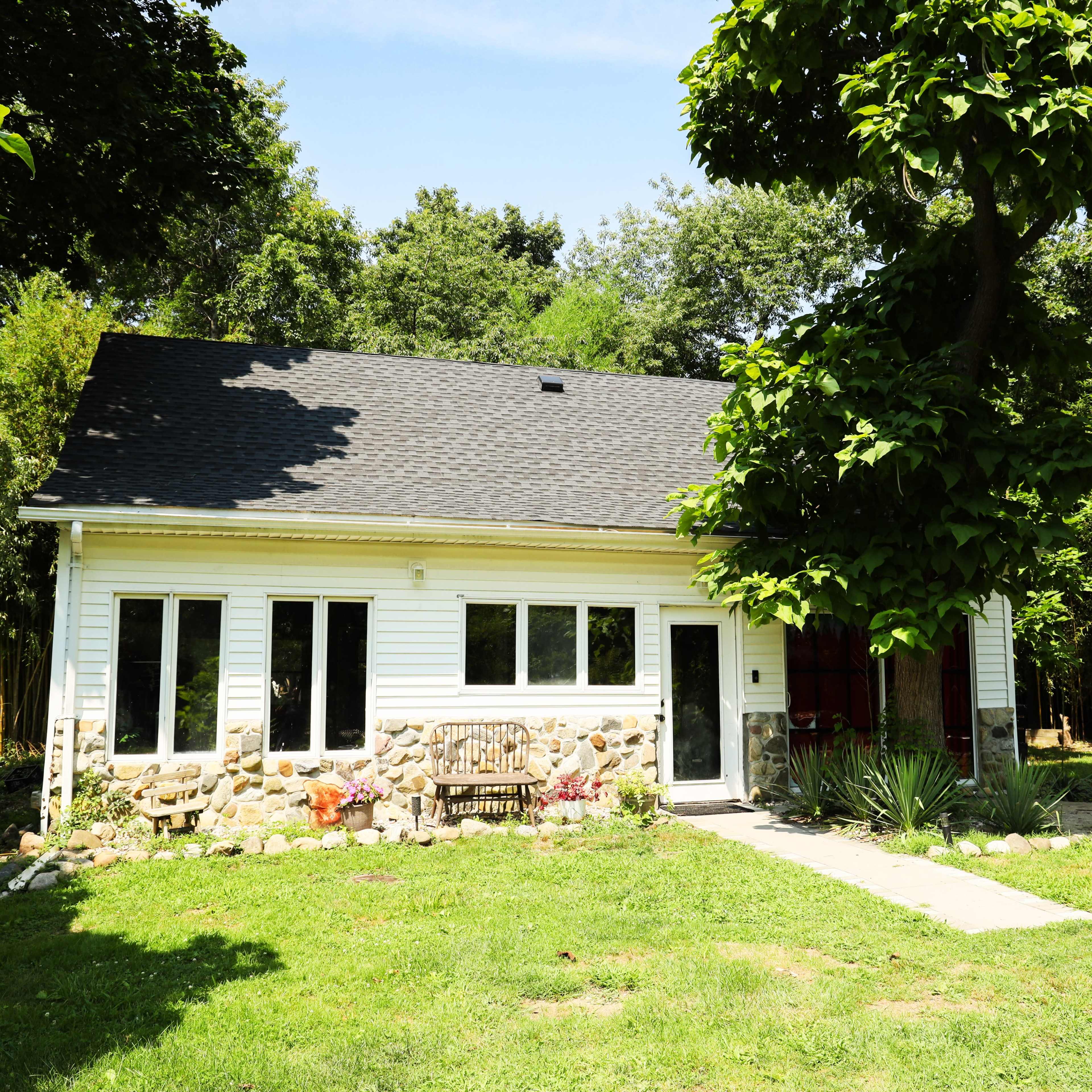 A single-story house with a stone facade and a wooden bench sits in a grassy area surrounded by trees.
