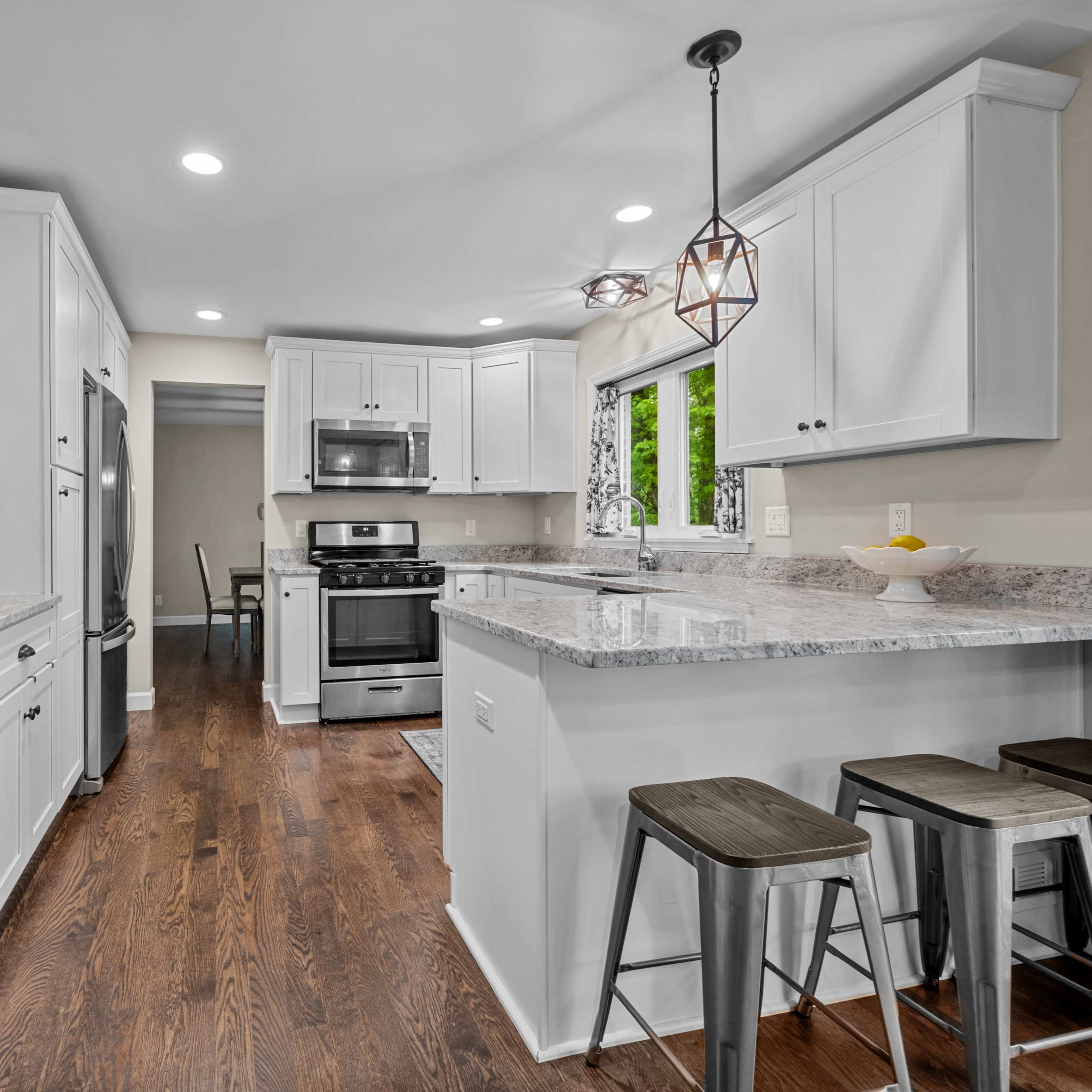 A modern kitchen featuring white cabinetry, granite countertops, stainless steel appliances, and a breakfast bar with three metal stools.
