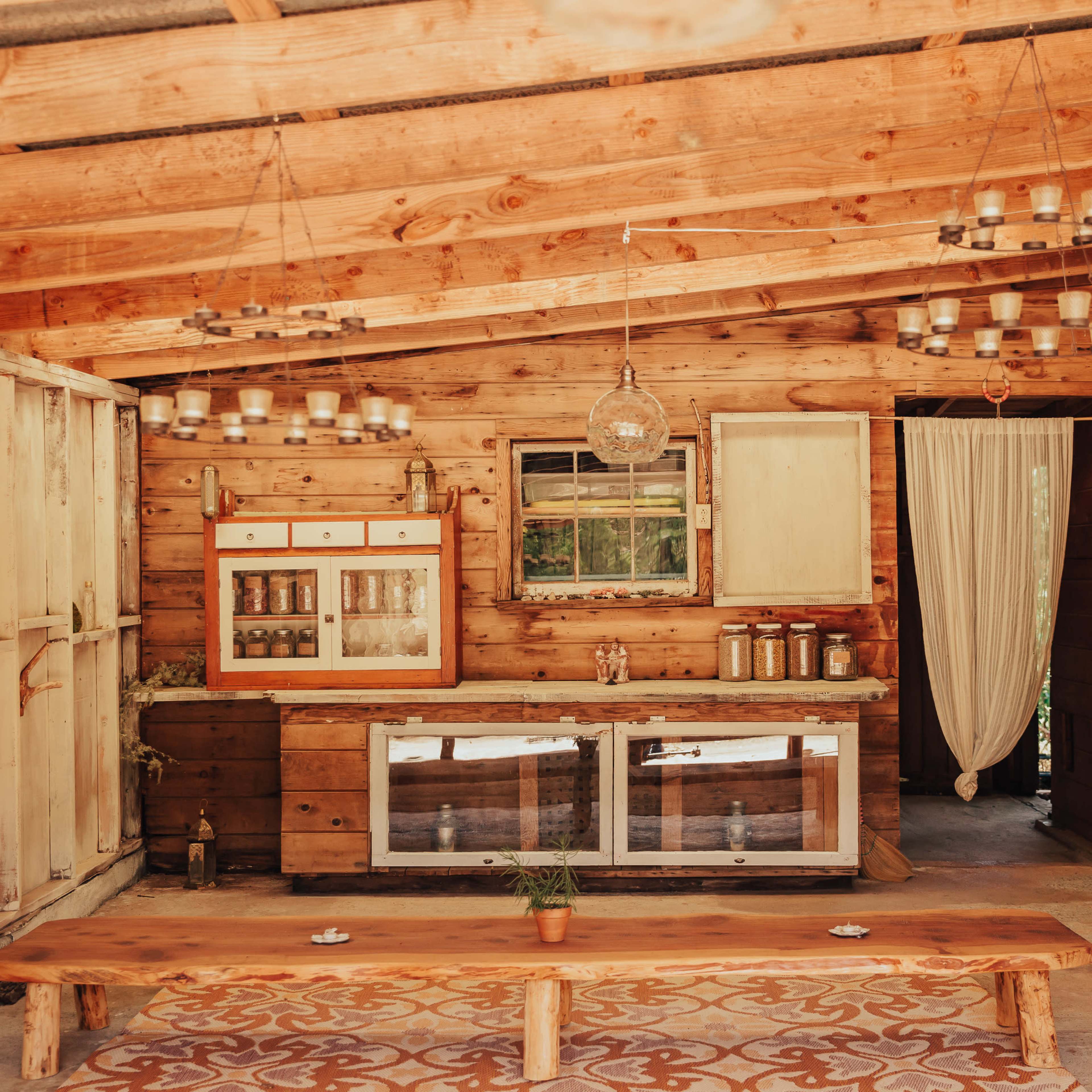 A wooden interior features a rustic kitchen area with a countertop, glass-fronted cabinets, chandeliers, and a small table with a plant on it.