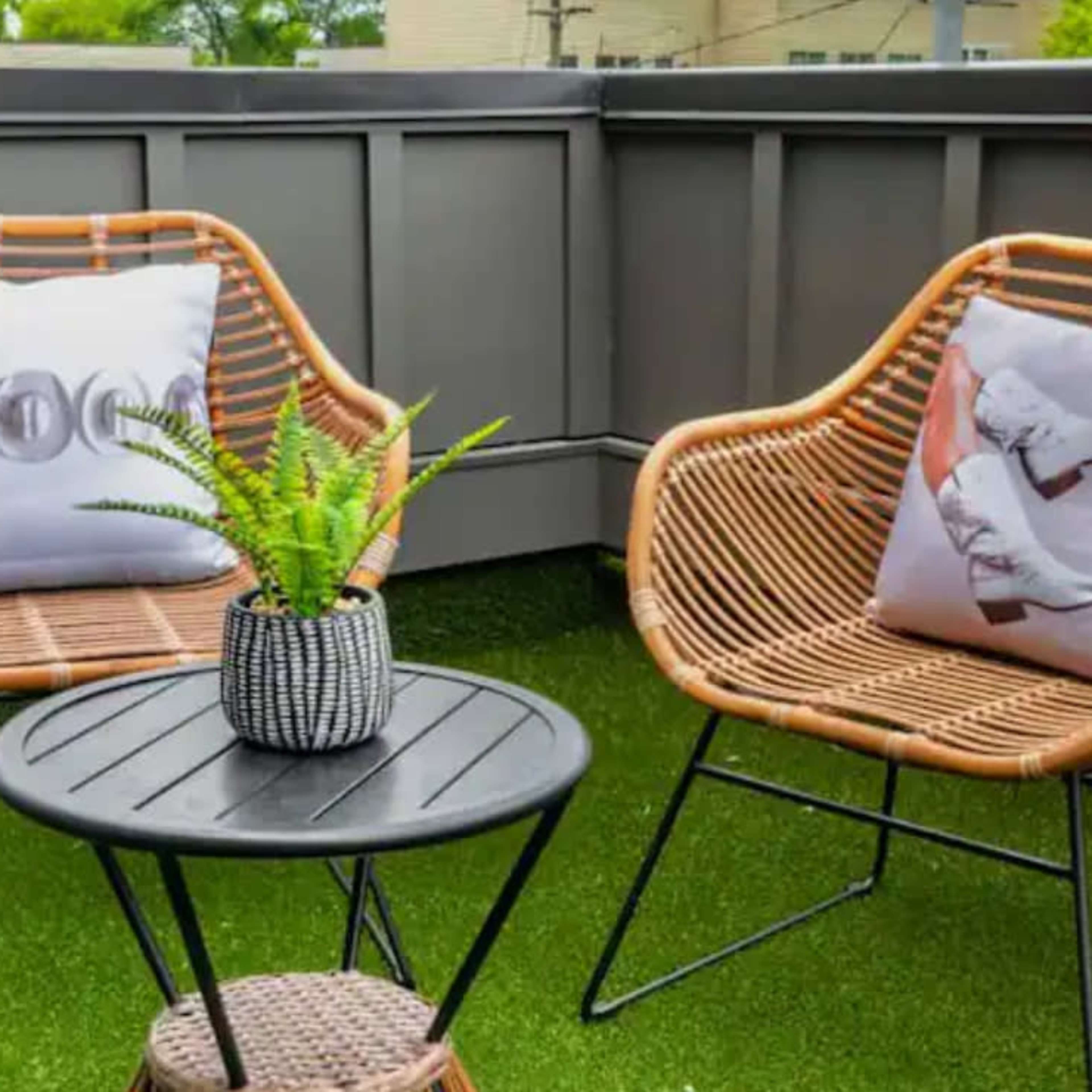 Two rattan chairs with decorative pillows facing a small black table, set on a grassy balcony.