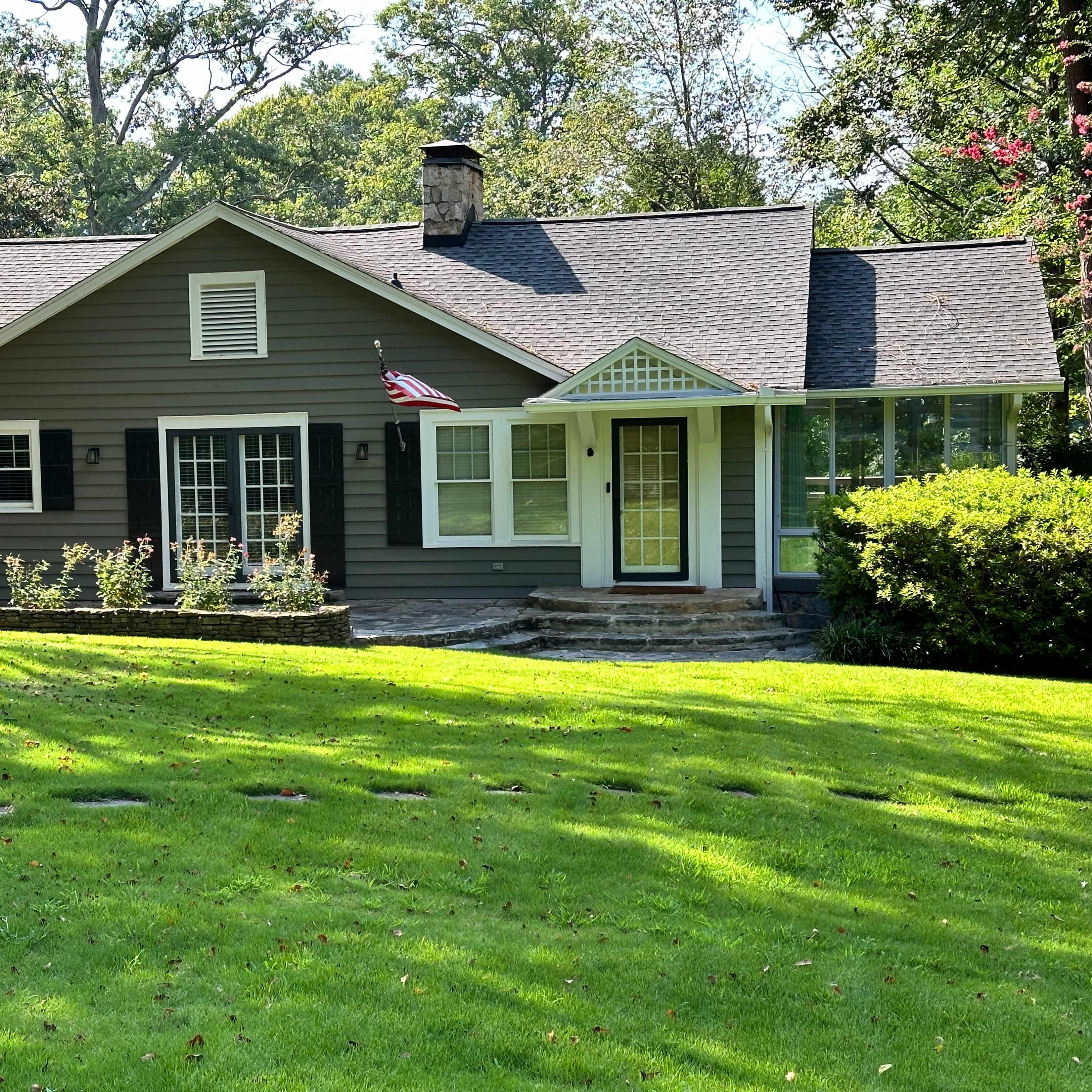 A single-story, gray house with a front porch and an American flag, surrounded by a lawn and trees.