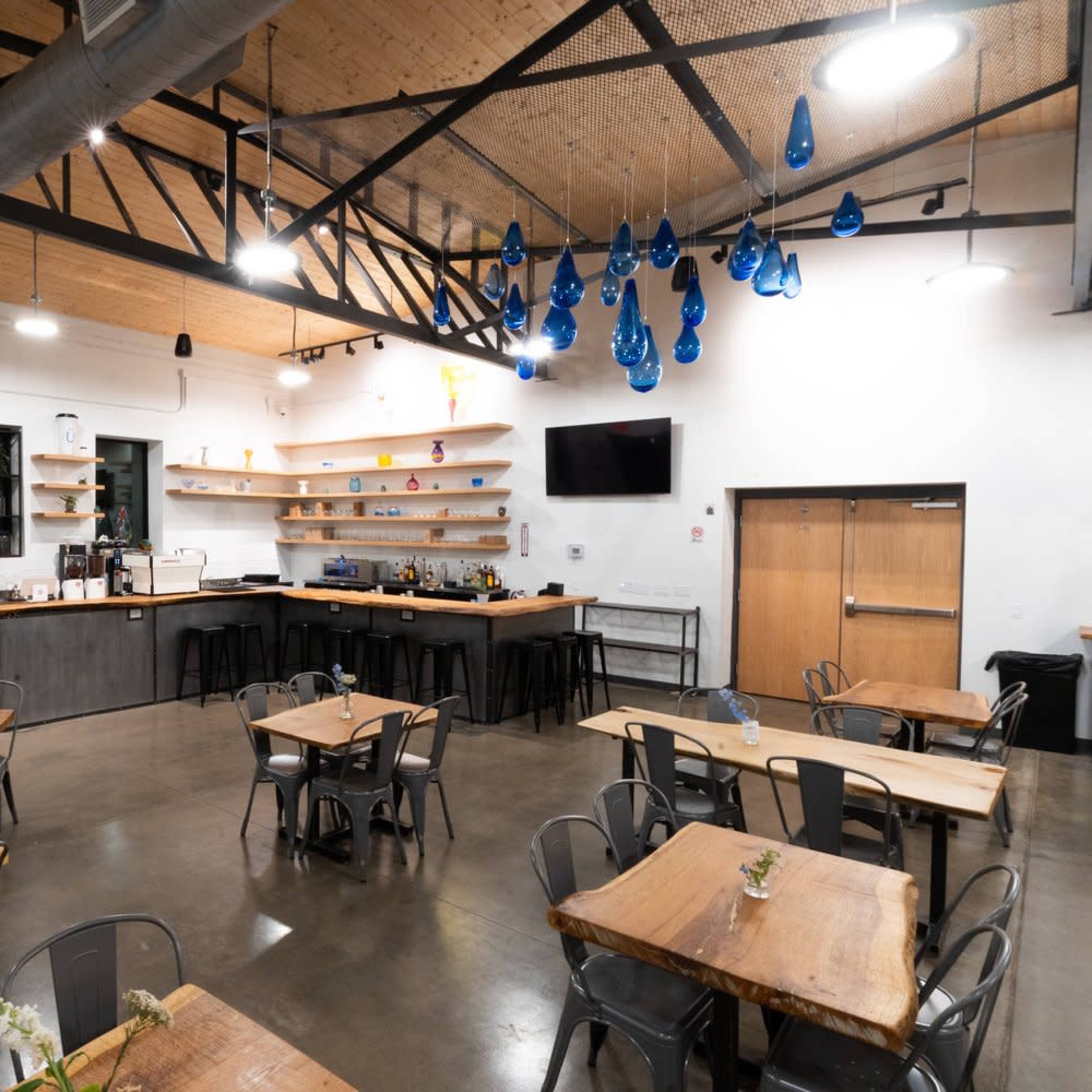 A modern café interior featuring wooden tables, metal chairs, and a bar area with shelves and blue decorative hanging lights.