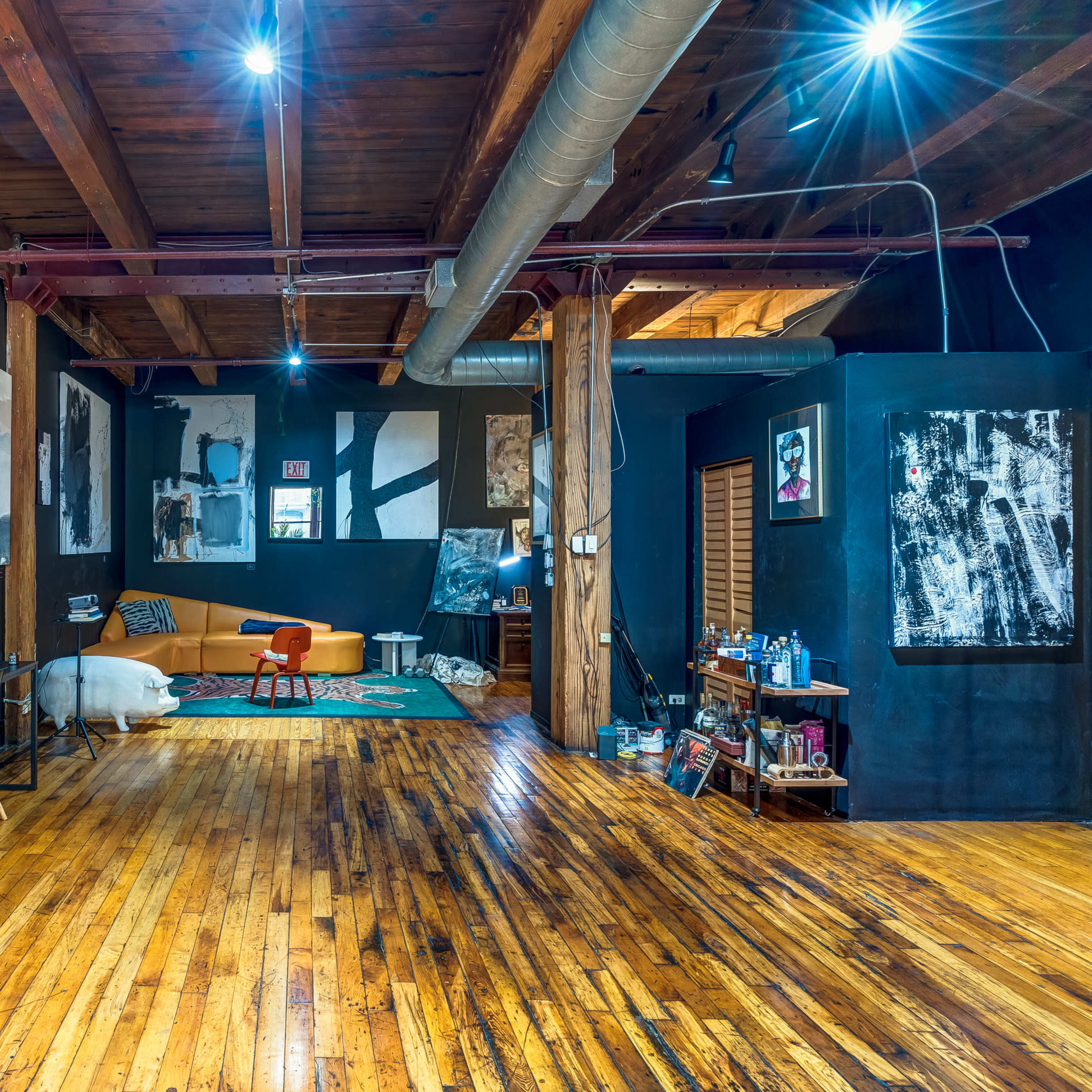 A spacious room with wooden floors, exposed wooden beams, and several abstract art pieces on dark walls, along with a couch and a dining table set against the backdrop.