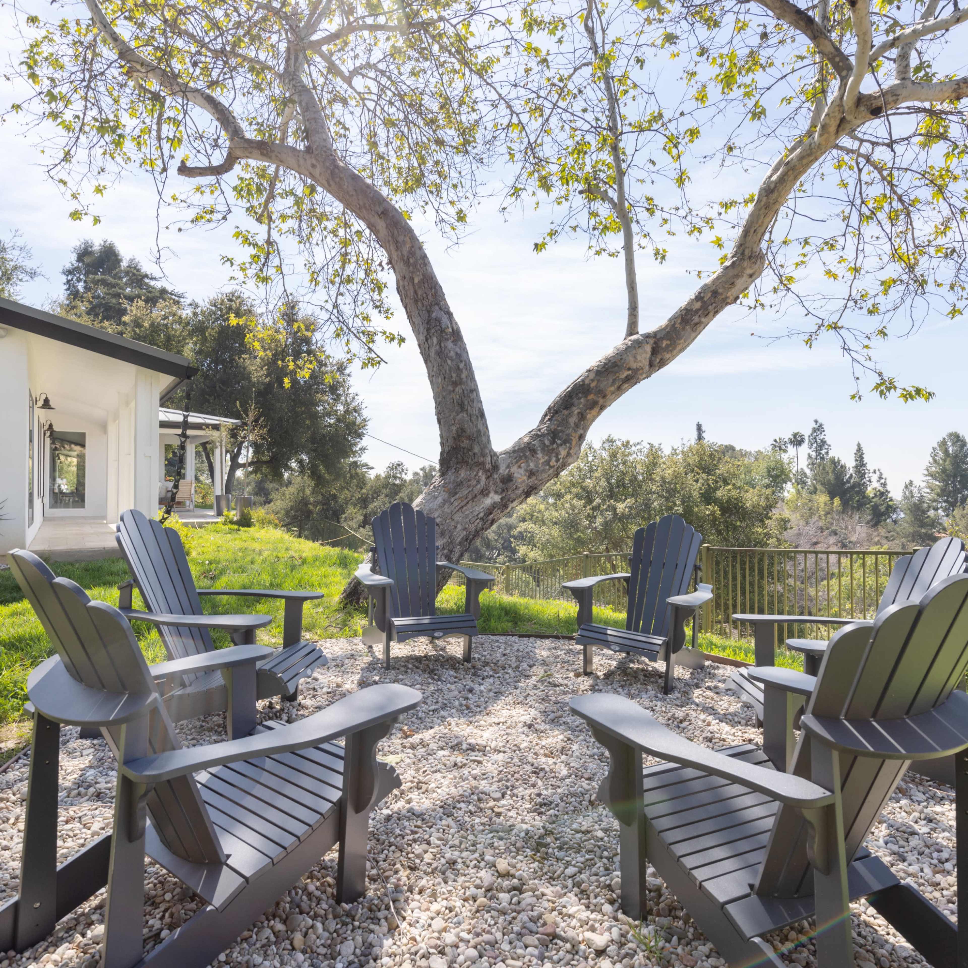 A circular seating area with black Adirondack chairs is surrounded by gravel and overlooks a green landscape and a large tree.